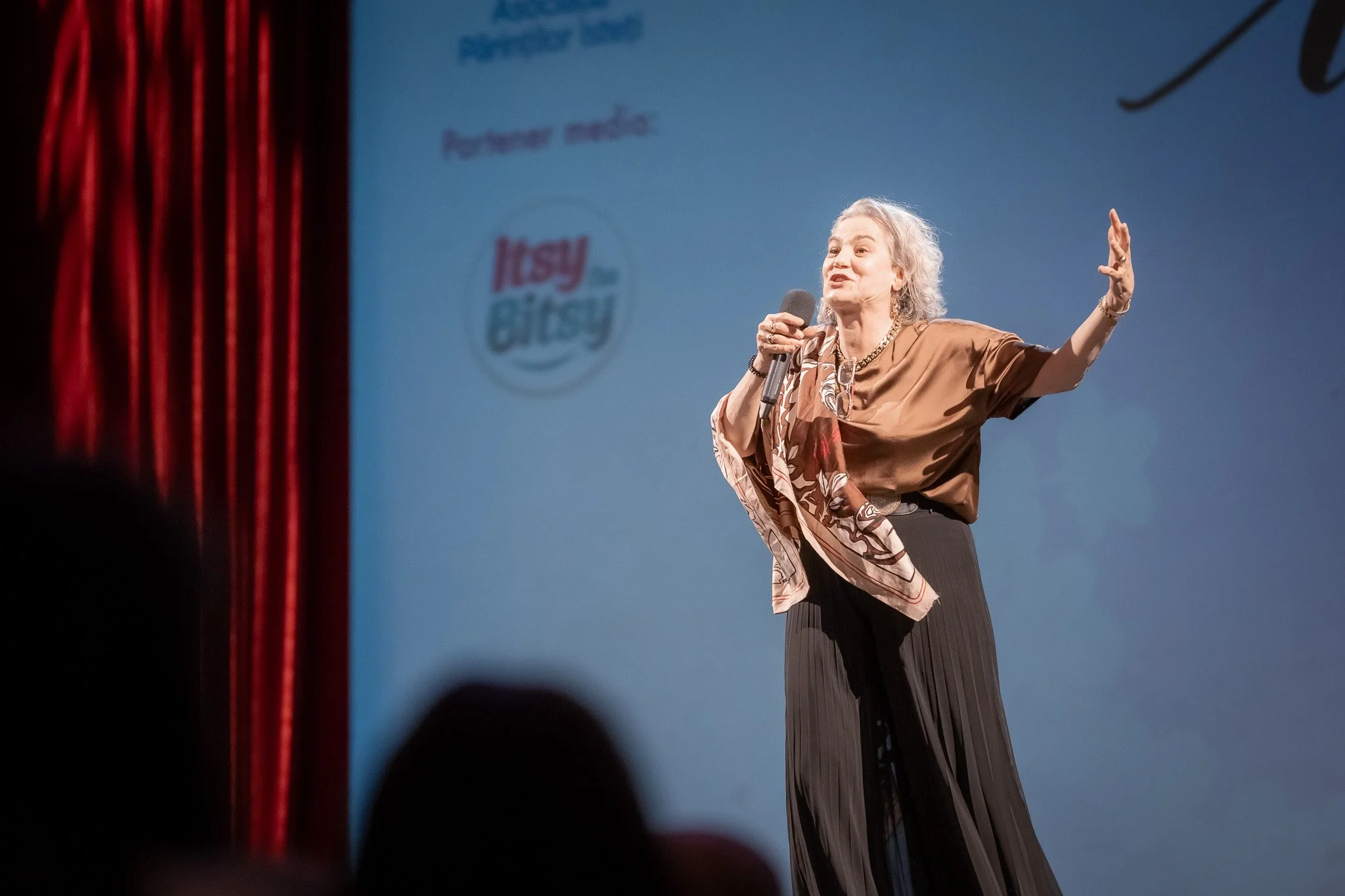 An older woman with gray hair speaking into a microphone on stage, with a presentation screen behind her displaying logos and text.