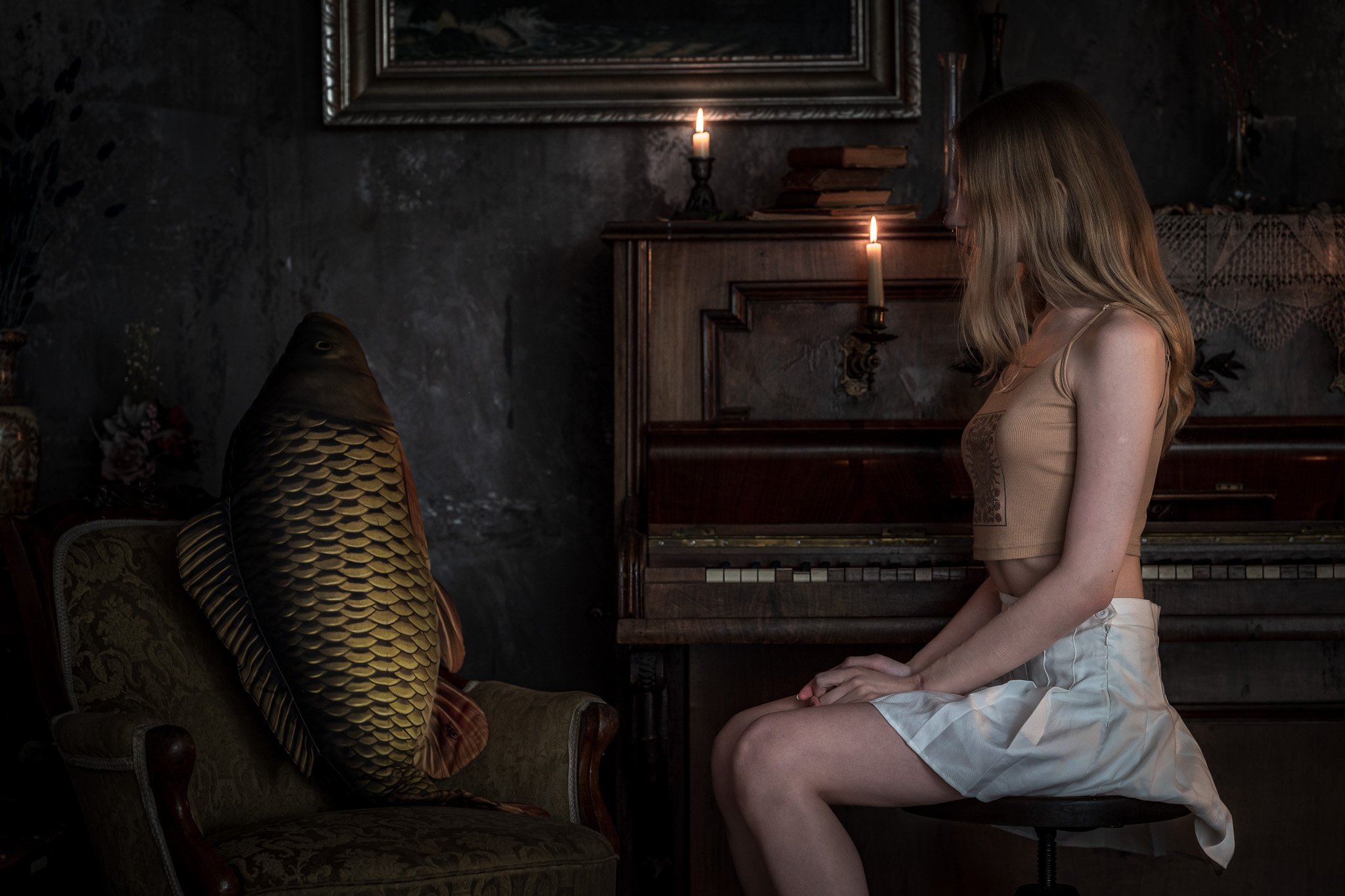 A woman with long blonde hair sitting on a stool in front of a vintage piano, looking at a large fish sitting on a sofa. The room is dimly lit by candles on top of the piano and a wall lamp, with dark walls and picture frames behind.