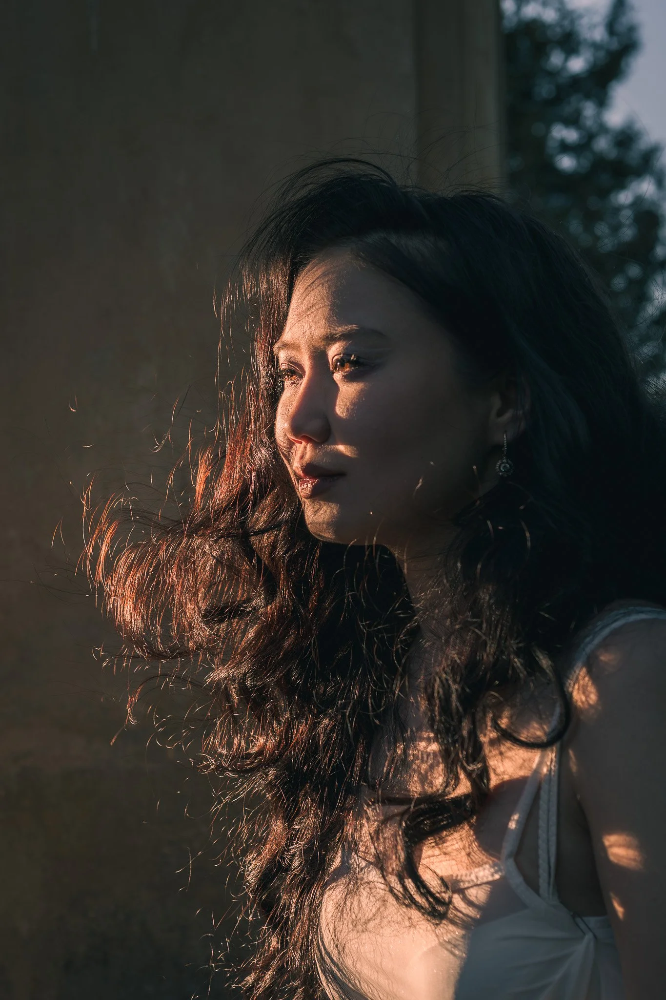 A woman with long, wavy dark hair looking slightly downward and to her left, illuminated by warm sunlight. She is wearing earrings and a white top, standing outdoors with a blurred background of trees.