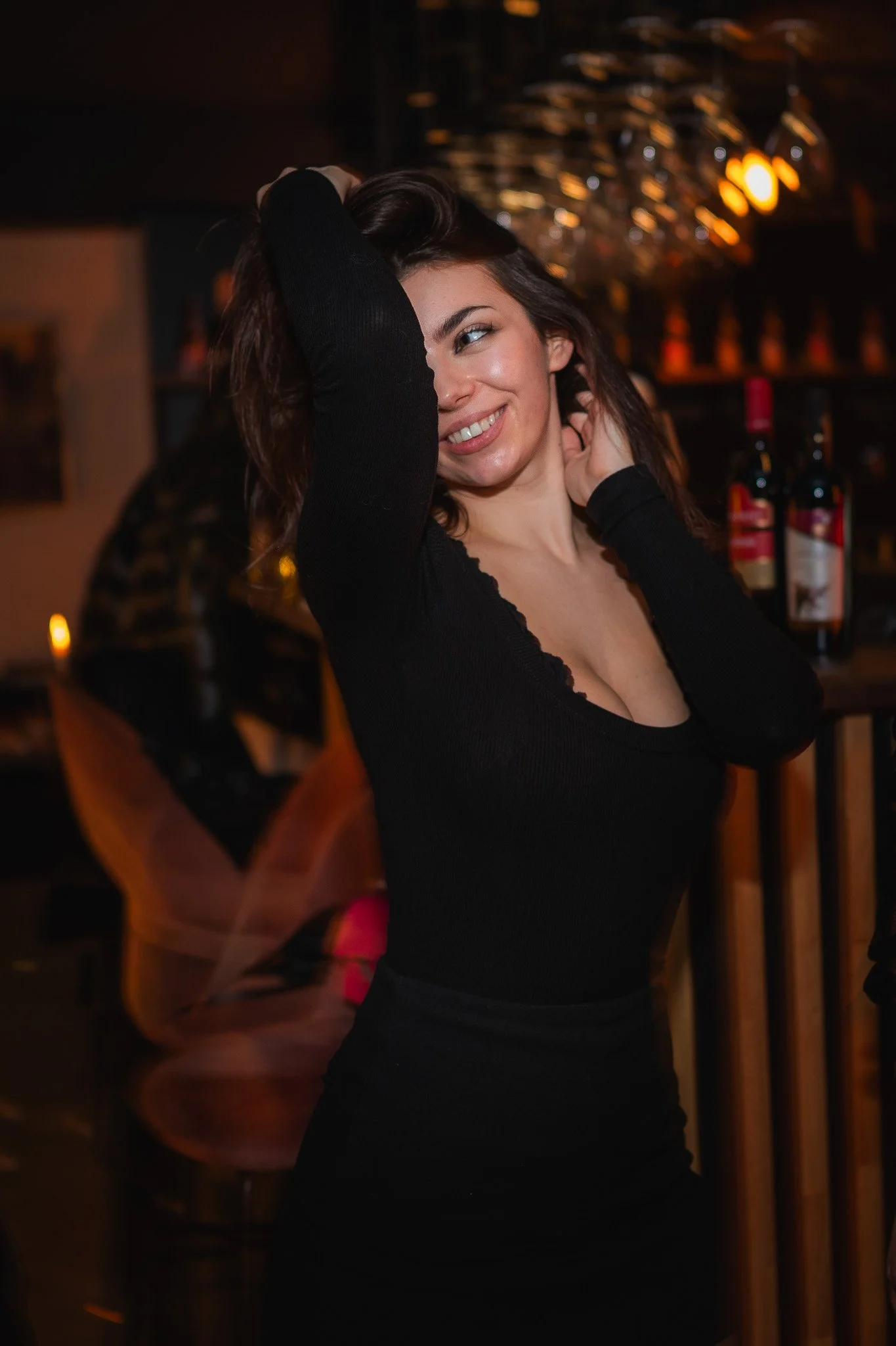 A woman smiling in a dimly lit bar or restaurant, wearing a black long-sleeved top, with bottles of wine on the shelf behind her. Fotograf Praha