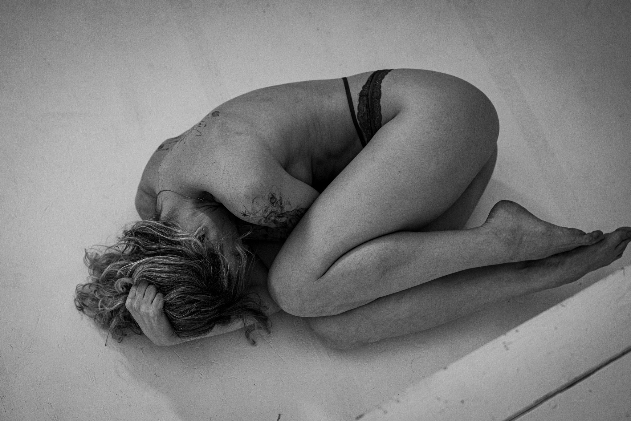 Black and white photo of a person curled up on the floor in a fetal position, wearing underwear, with visible tattoos.