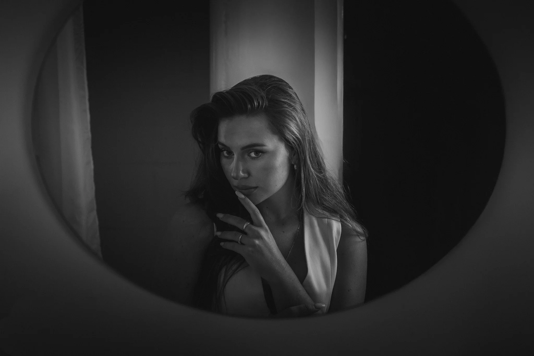 Černobílá fotografie ženy dívající se do zrcadla, ve které je vidět její odraz. Má dlouhé vlasy a jedním prstem se dotýká rtů. Vyrobeno v Praze.