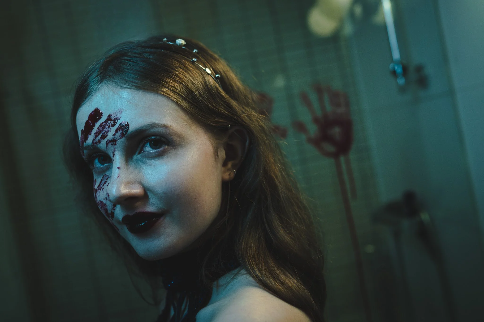 A young woman with dark hair and face paint resembling a skull, gazing into the distance with an intense expression in a dimly lit room featuring a showerhead and red graffiti on the wall.