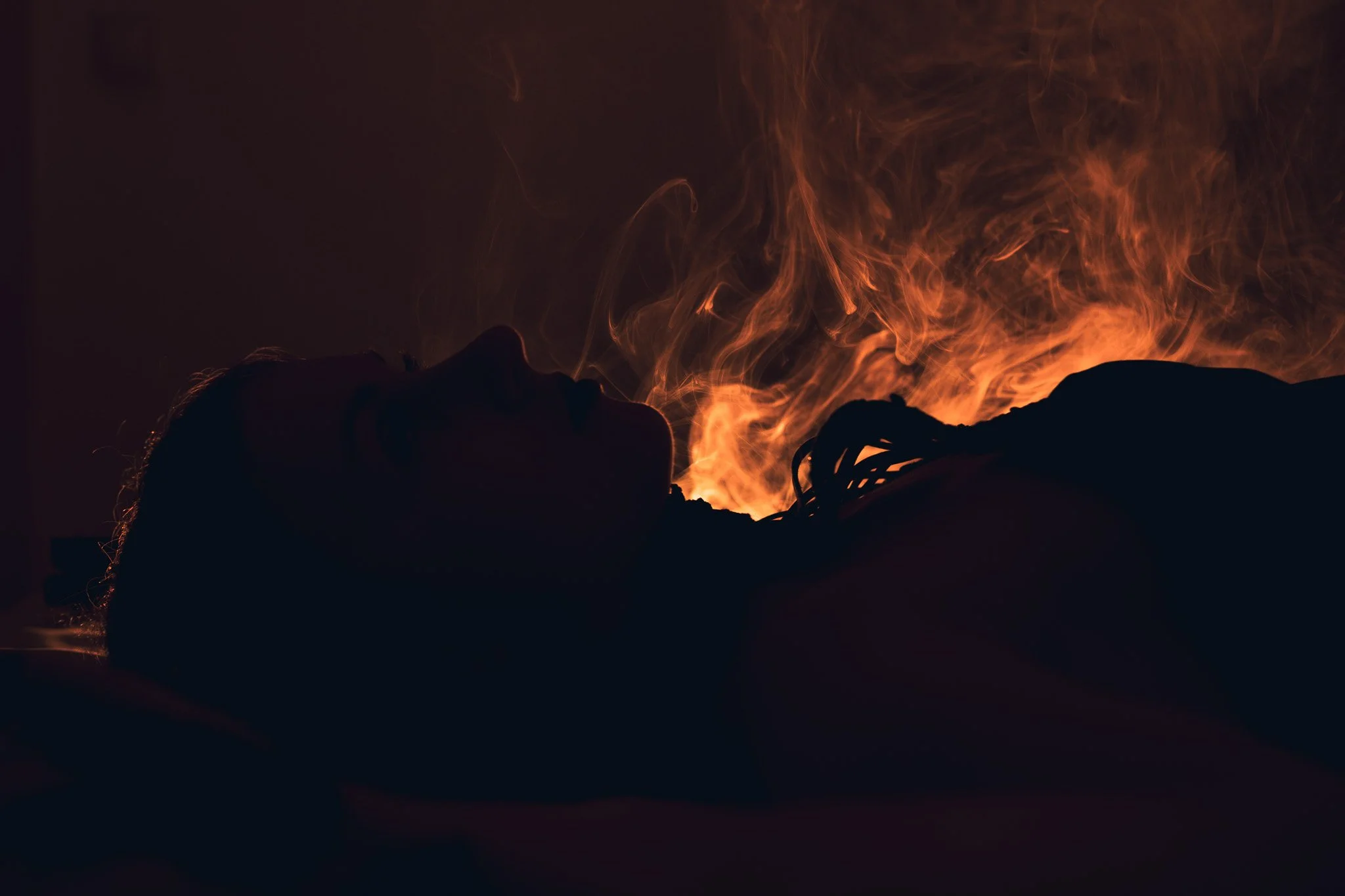 A person lying down with flames surrounding their head and face against a dark background.