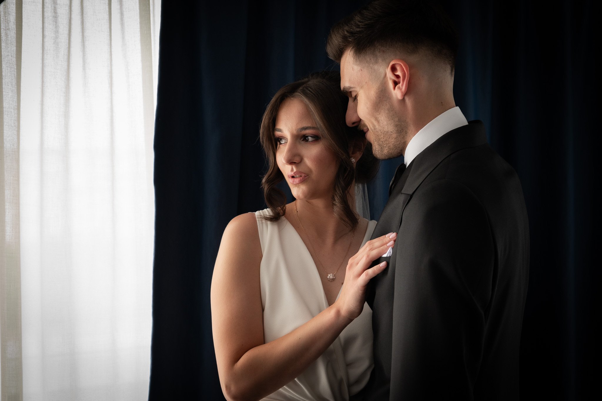 A bride and groom in a tender moment, standing close with the bride touching the groom's chest, indoors near a window with white curtains and navy blue drapes.