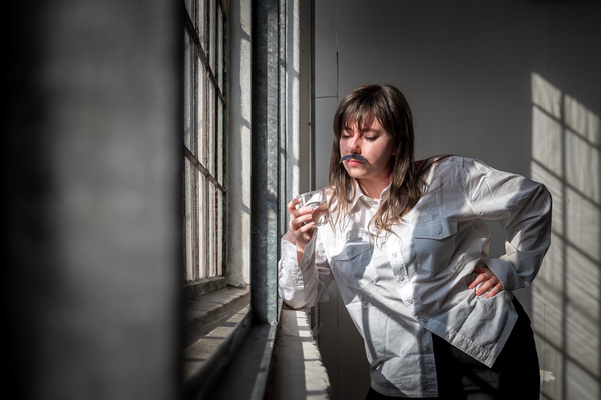 Person wearing a white shirt and fake mustache, holding a glass, standing by a window.