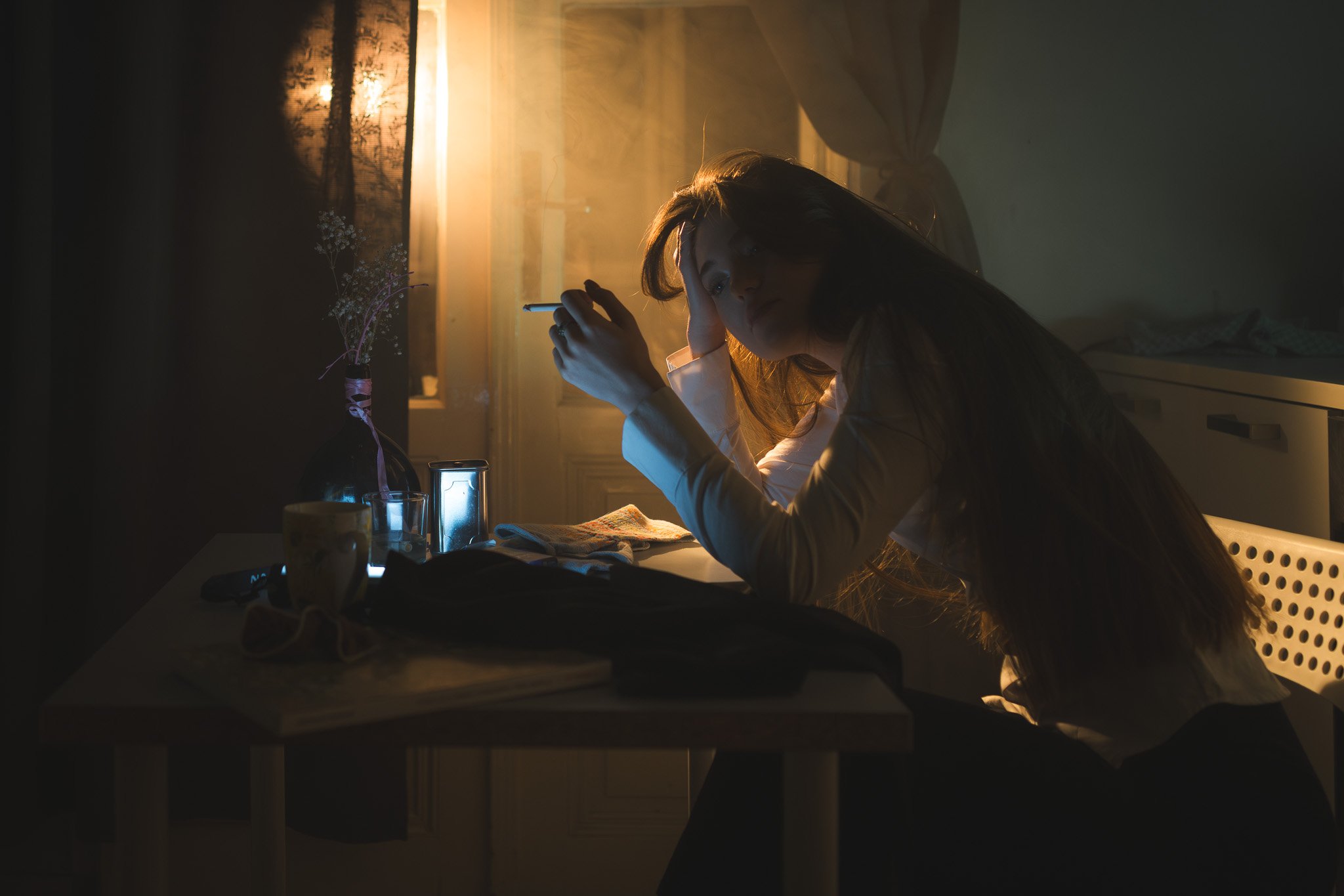 A photograph of a woman with long hair sitting at a table in a dimly lit room, bathed in warm light streaming in from the window. 