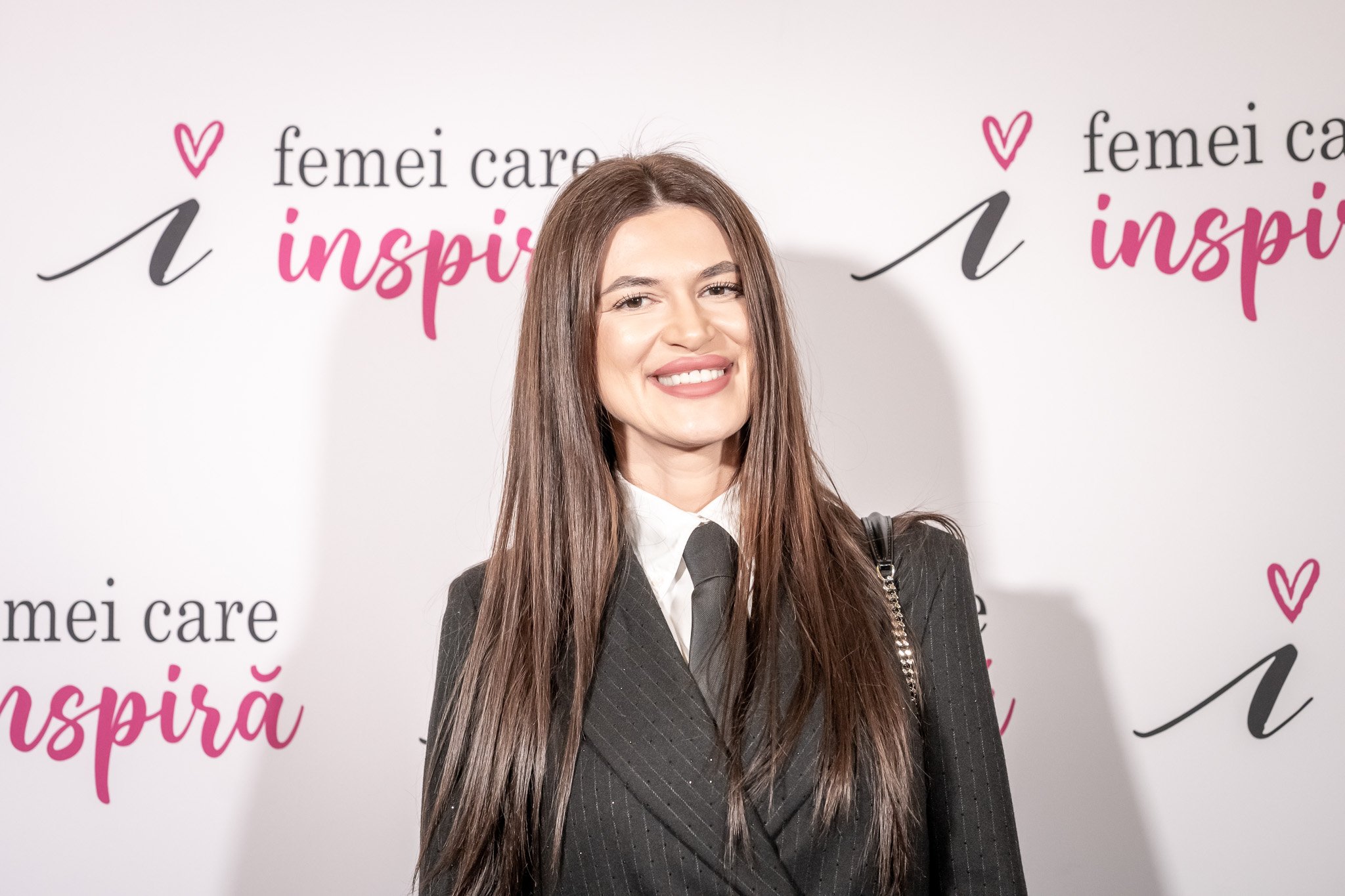 A smiling woman with long brown hair, wearing a black pinstripe blazer, a white shirt, and a black tie, standing in front of a white backdrop with pink and black text that reads "women who inspire."