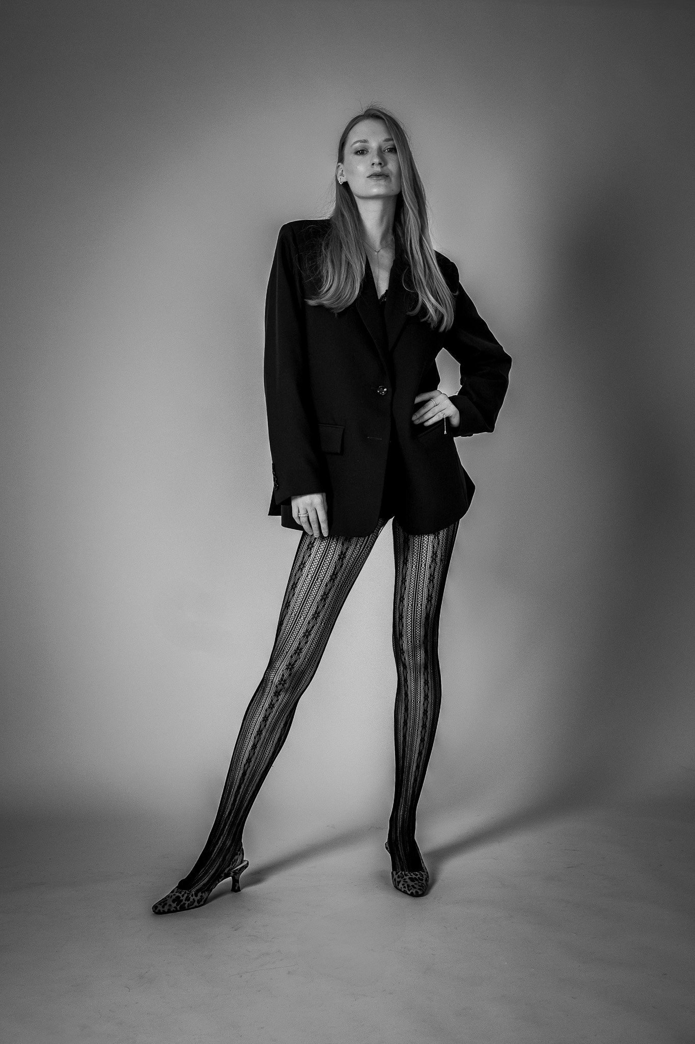 Black and white photo of a person wearing a black blazer, patterned tights, and leopard print heels, standing in a pose against a plain background.