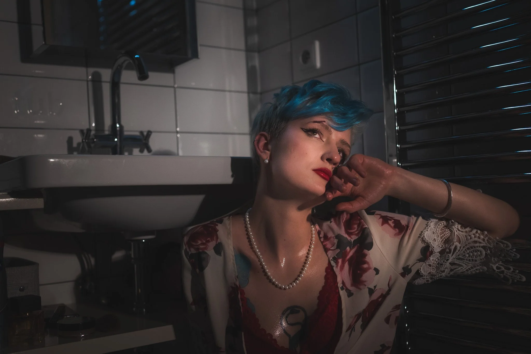 A woman with short blue hair and red lipstick leaning against the wall in a dimly lit bathroom, wearing a floral robe, pearl necklace, and red lingerie.