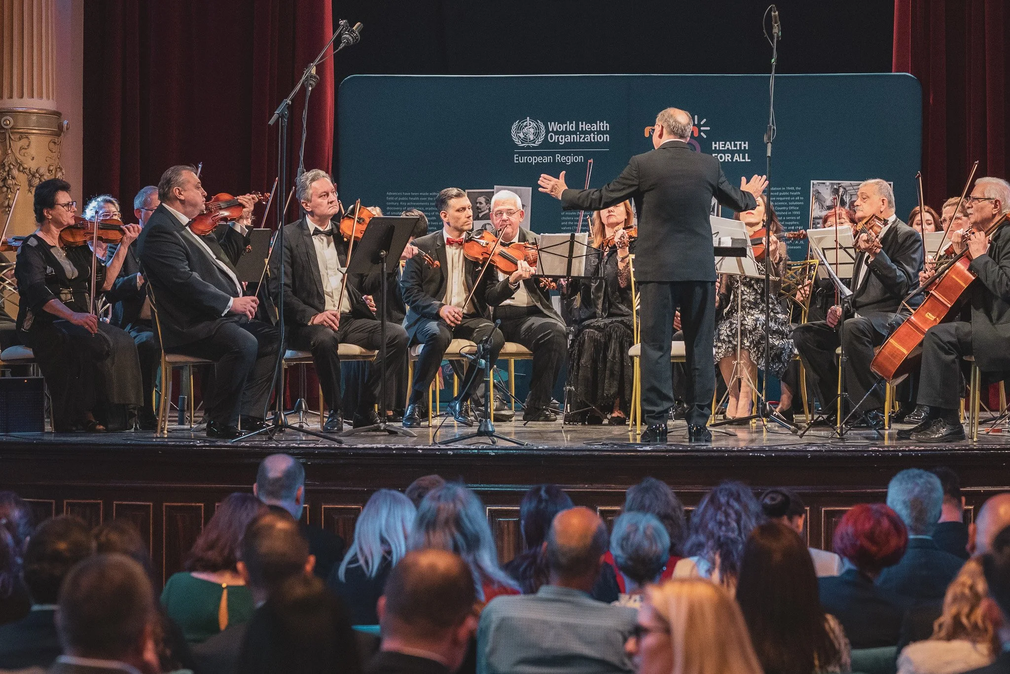 An orchestra performing on stage with a conductor leading them, in front of an audience. The backdrop has the World Health Organization logo and text.