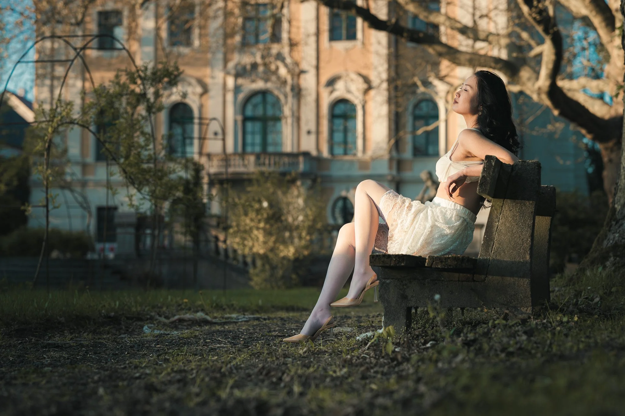 A woman sitting on a park bench with her eyes closed and face tilted upwards, enjoying the sunlight, in front of a large historic building with trees around.