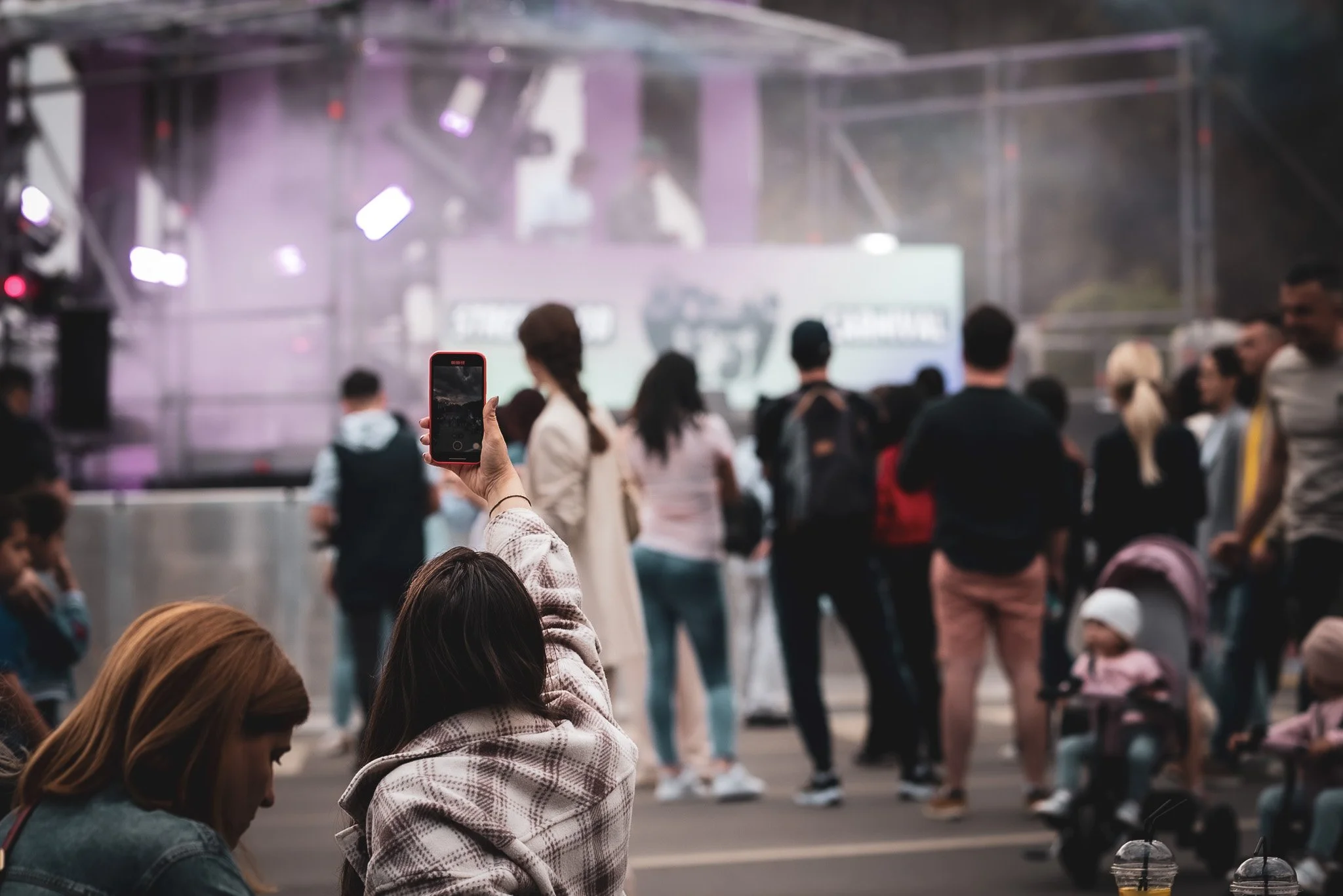 People gathered in front of a stage at an outdoor event, some looking at their phones, with stage lights and a screen in the background. Fotograf Praha