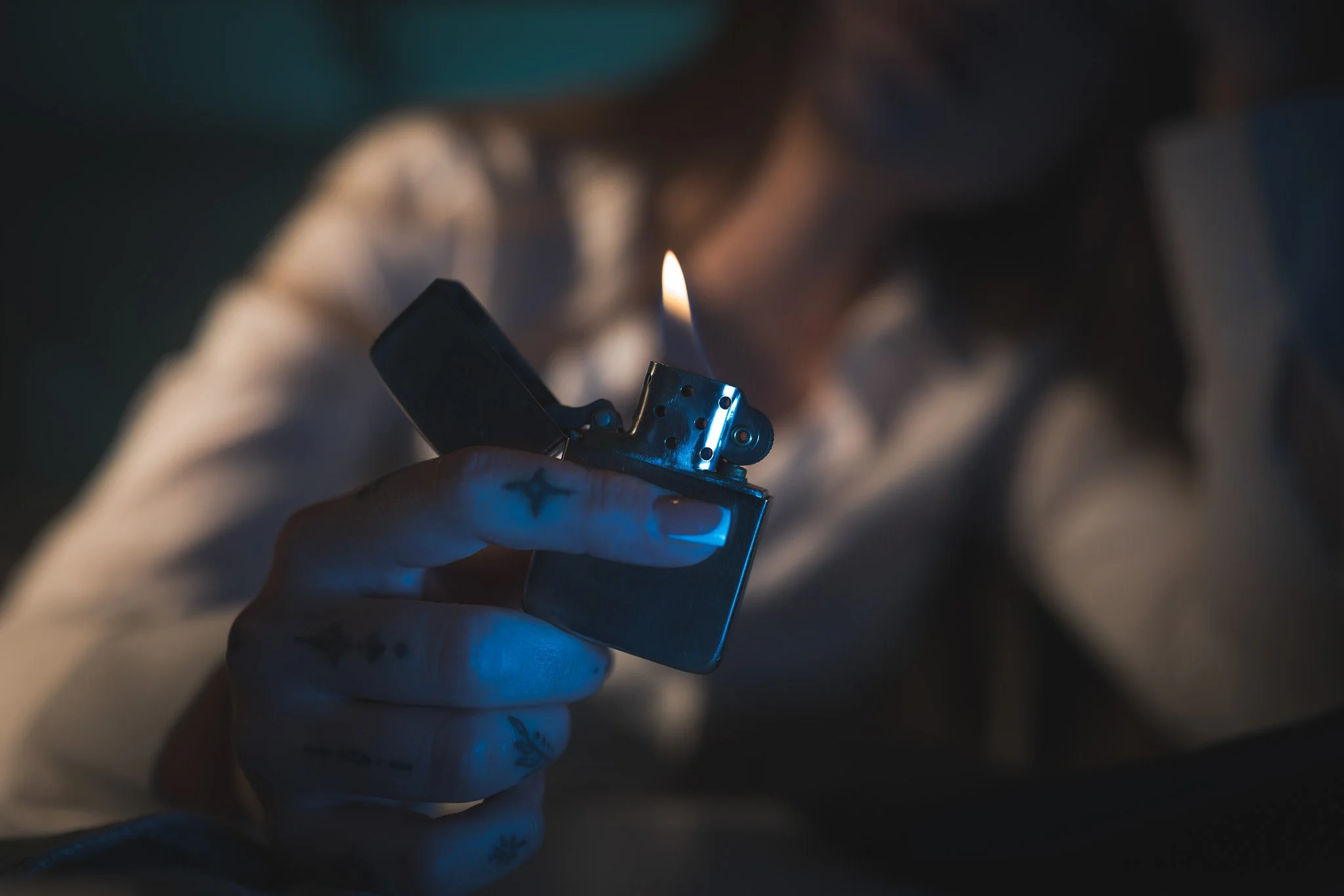 A person is holding a lit Zippo-style lighter with a flame, wearing glasses and a white shirt, in a dimly lit room.