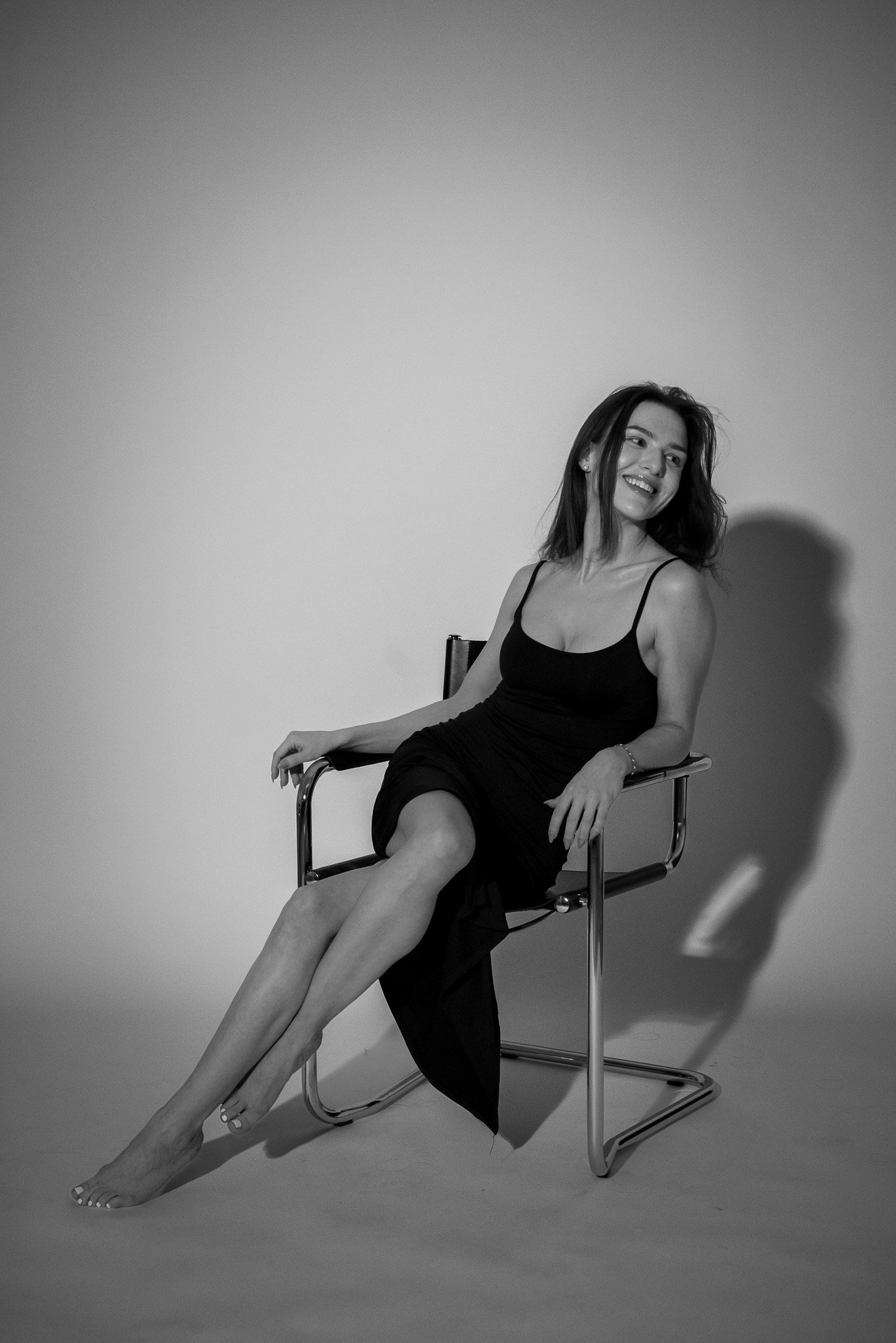 Black and white photo of a woman sitting on a chair, smiling and wearing a sleeveless dress.
