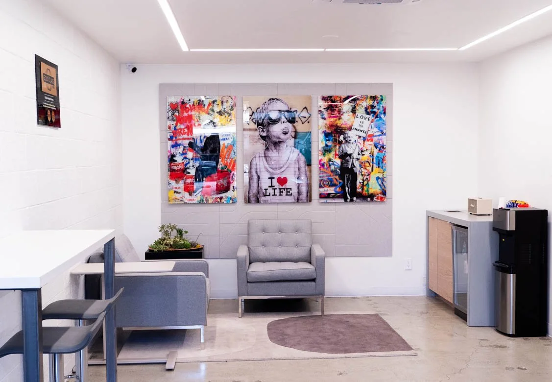 Modern lounge area with three colorful graffiti-style art posters on the wall, gray armchair, small gray sofa, potted plant, water dispenser, mini fridge, and a countertop with paper cups.