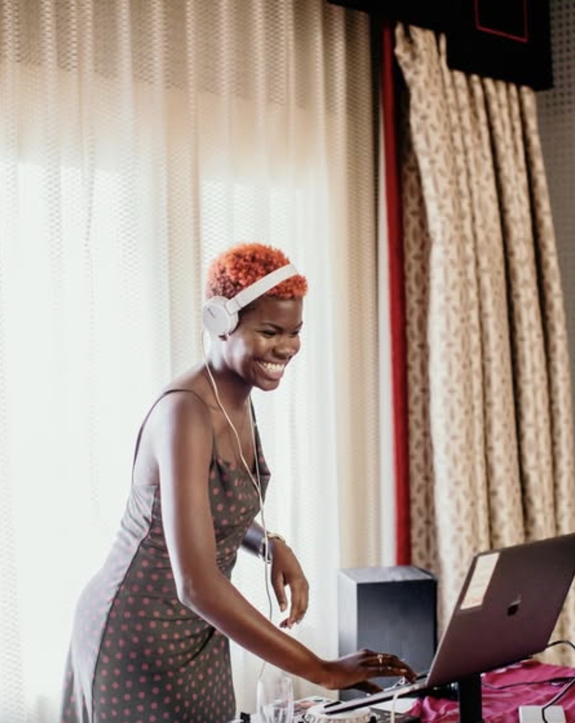 Woman with short red hair smiling while using a laptop and wearing white headphones in a room with cream curtains and patterned drapes.