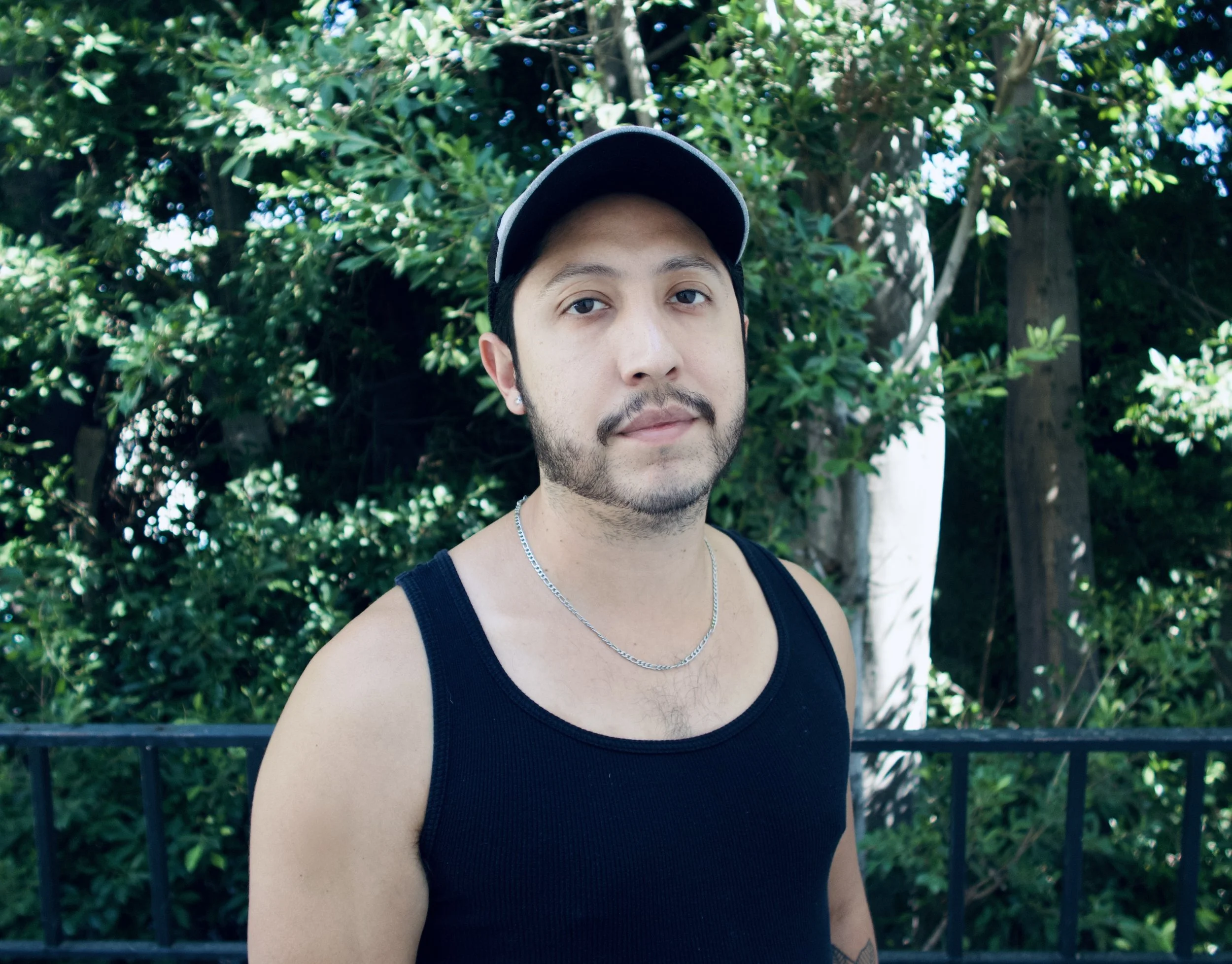 A young man wearing a black tank top, a silver chain, and a black baseball cap, standing outdoors in front of lush green trees and a black metal fence.