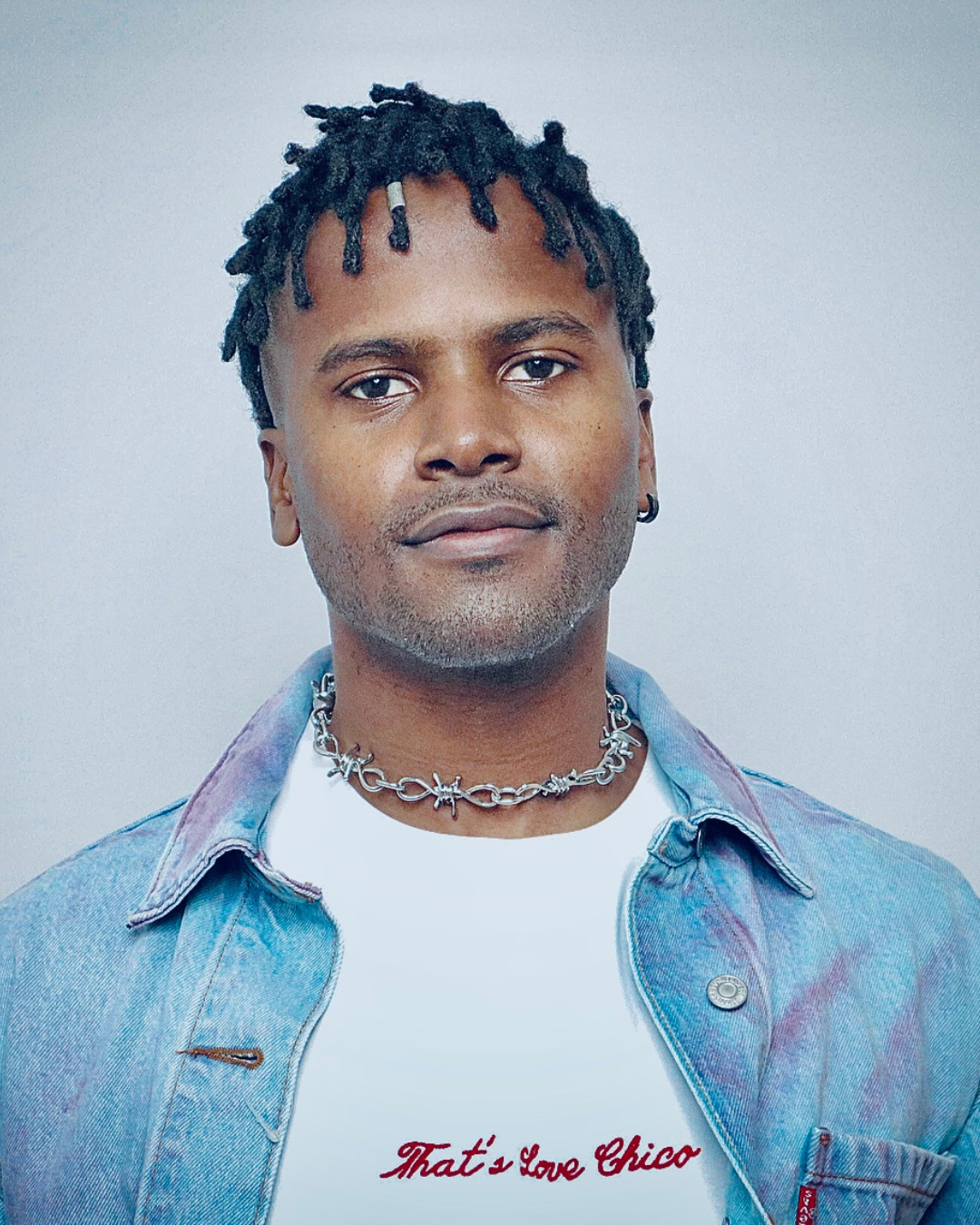A young man with short dreadlocks, wearing a silver chain necklace, a white t-shirt with red text, and a light denim jacket, looking directly at the camera with a neutral expression.