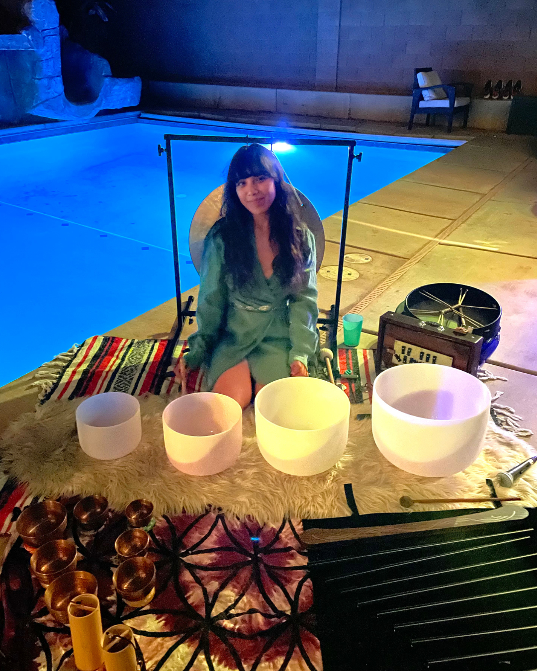 A woman sitting on a colorful rug by a pool, surrounded by singing bowls and musical instruments, illuminated by soft, multicolored lighting, suggesting a meditation or sound healing session.