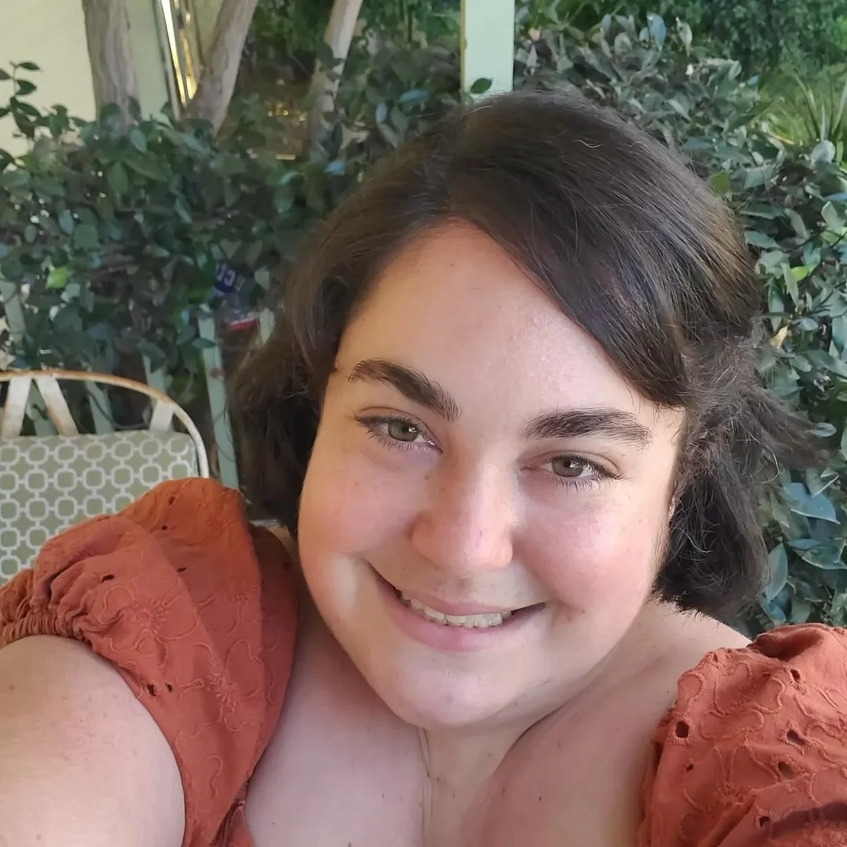 A woman with short dark hair and light eyes smiling outdoors with greenery in the background.