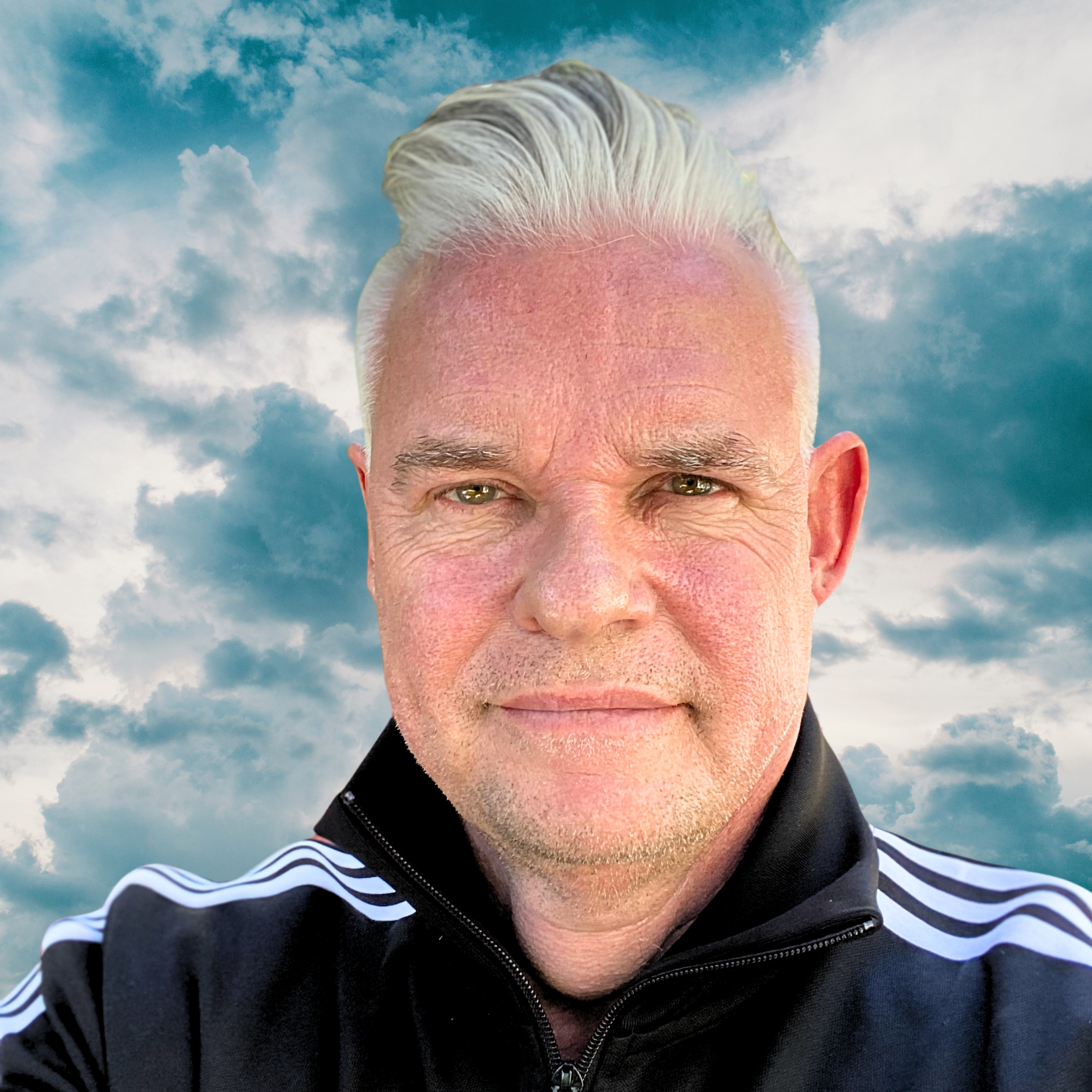 A man with light-colored hair and a slight stubble, wearing a black sports jacket with white stripes on the shoulders, standing outdoors with a background of a partly cloudy sky.