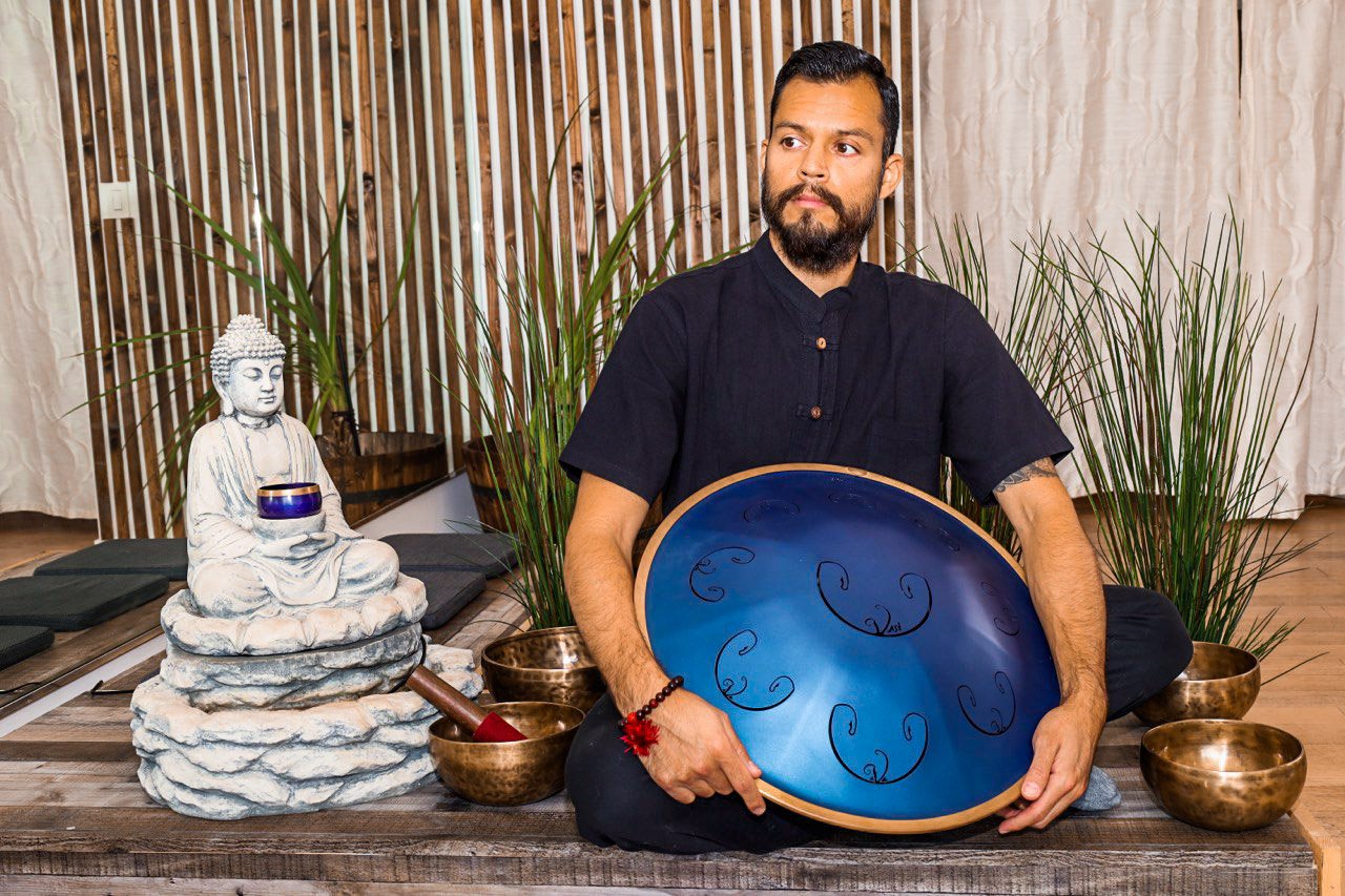 A man sitting cross-legged on a wooden platform, holding a large blue handpan, with a small Buddha statue, singing bowls, and green plants in the background.