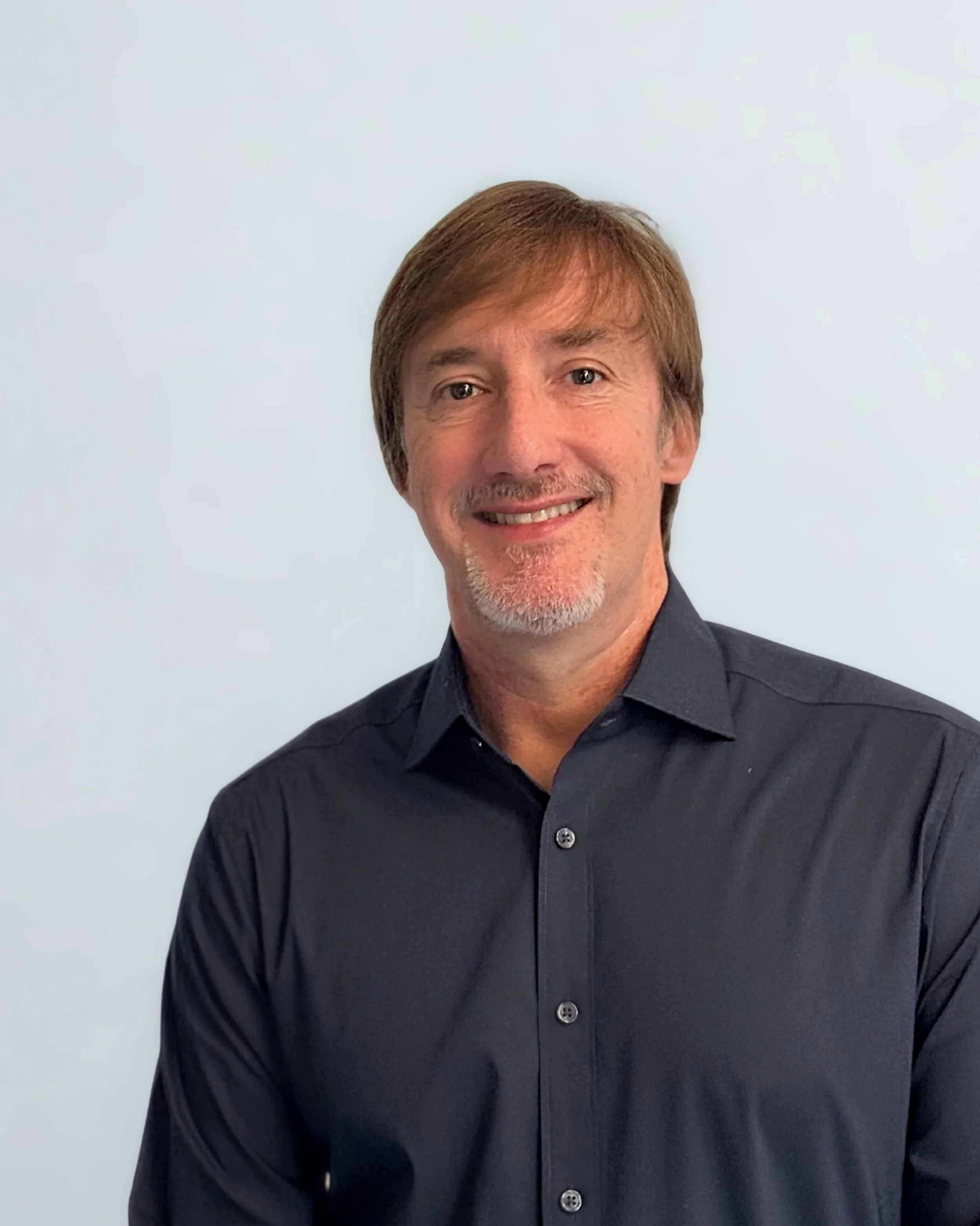 Portrait of a smiling man with brown hair, wearing a dark blue shirt, standing against a plain light background.