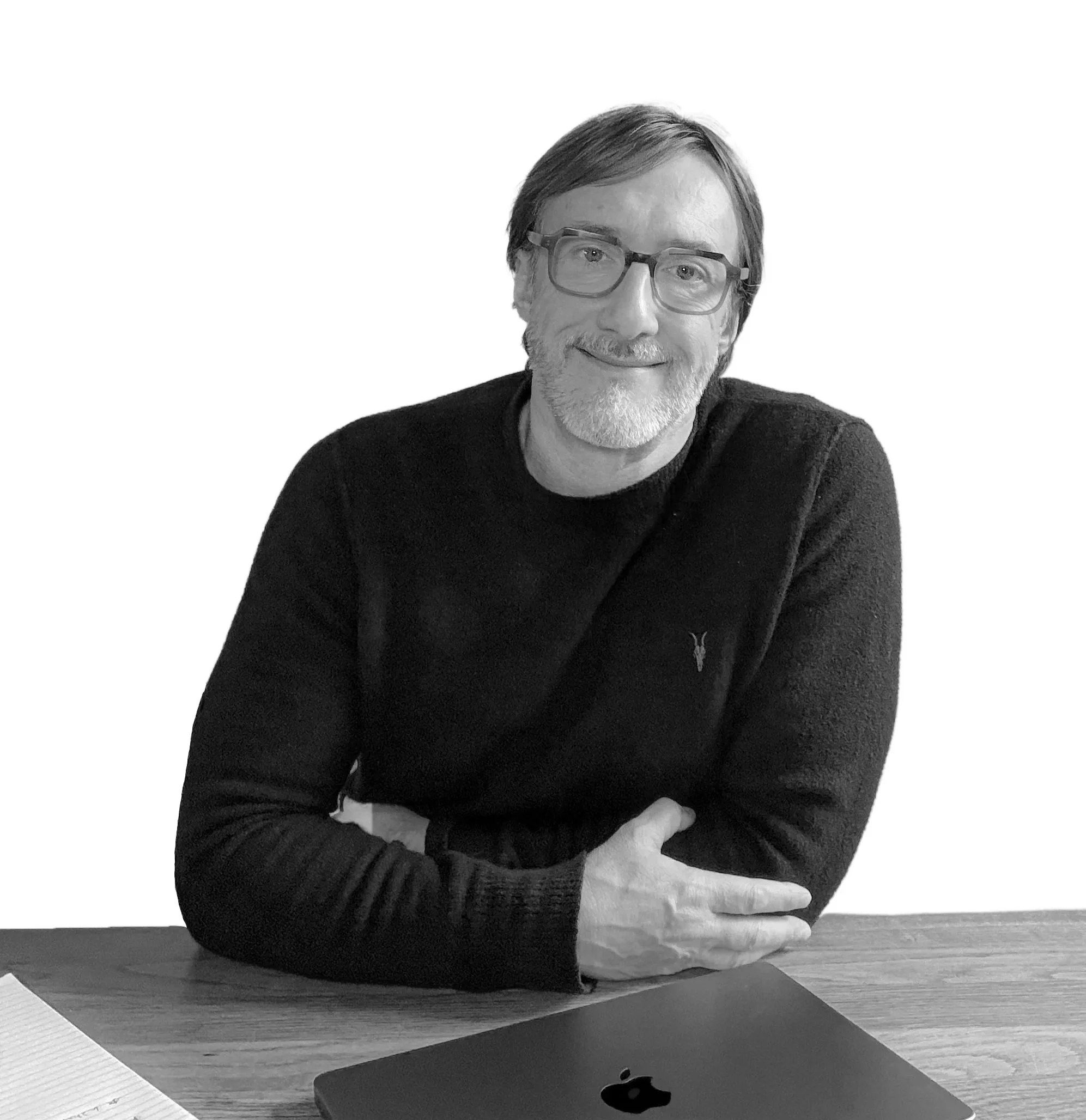A man with glasses and a beard smiling at the camera, sitting at a wooden table with a closed MacBook and some papers.