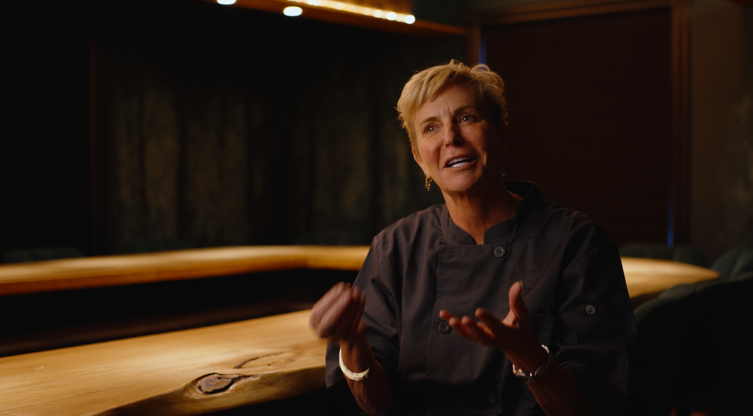 A woman in a black chef jacket is sitting at a wooden conference table in a dimly lit room, speaking with expressive hand gestures.