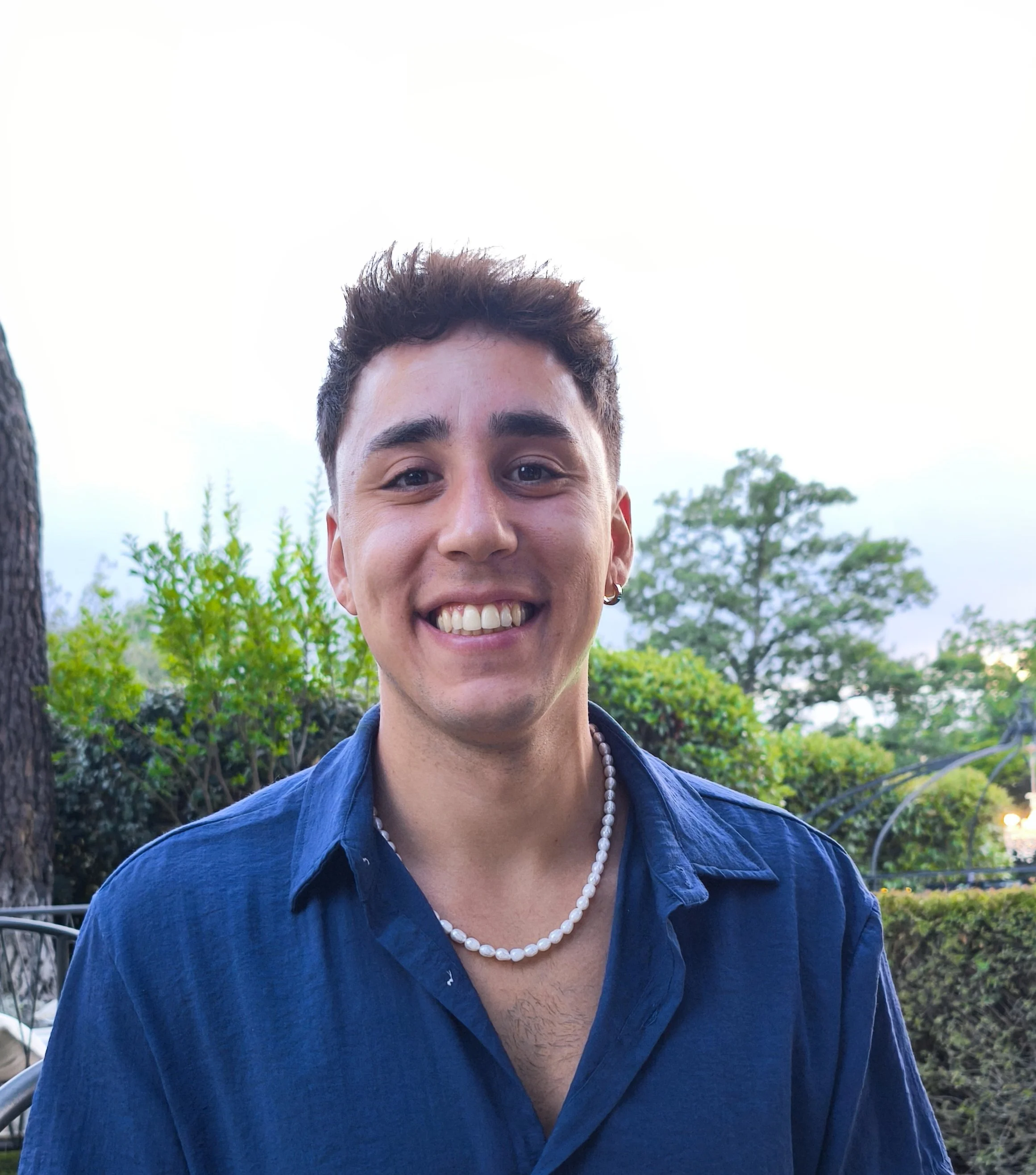 Ragazzo sorridente con camicia blu, collana di perle, orecchino, con sfondo di alberi e cielo sereno.