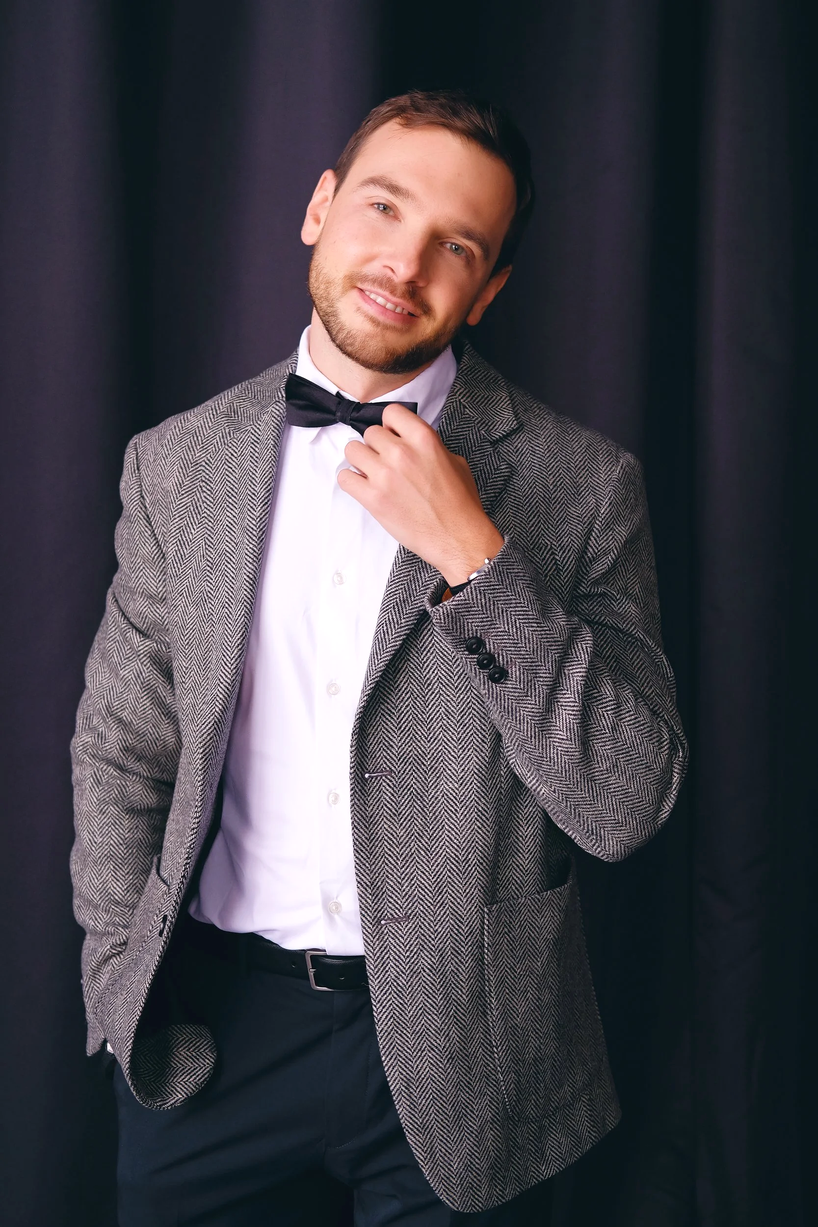 A man in a gray checkered blazer, white shirt, and black tie adjusting his tie, standing against a dark curtain background.