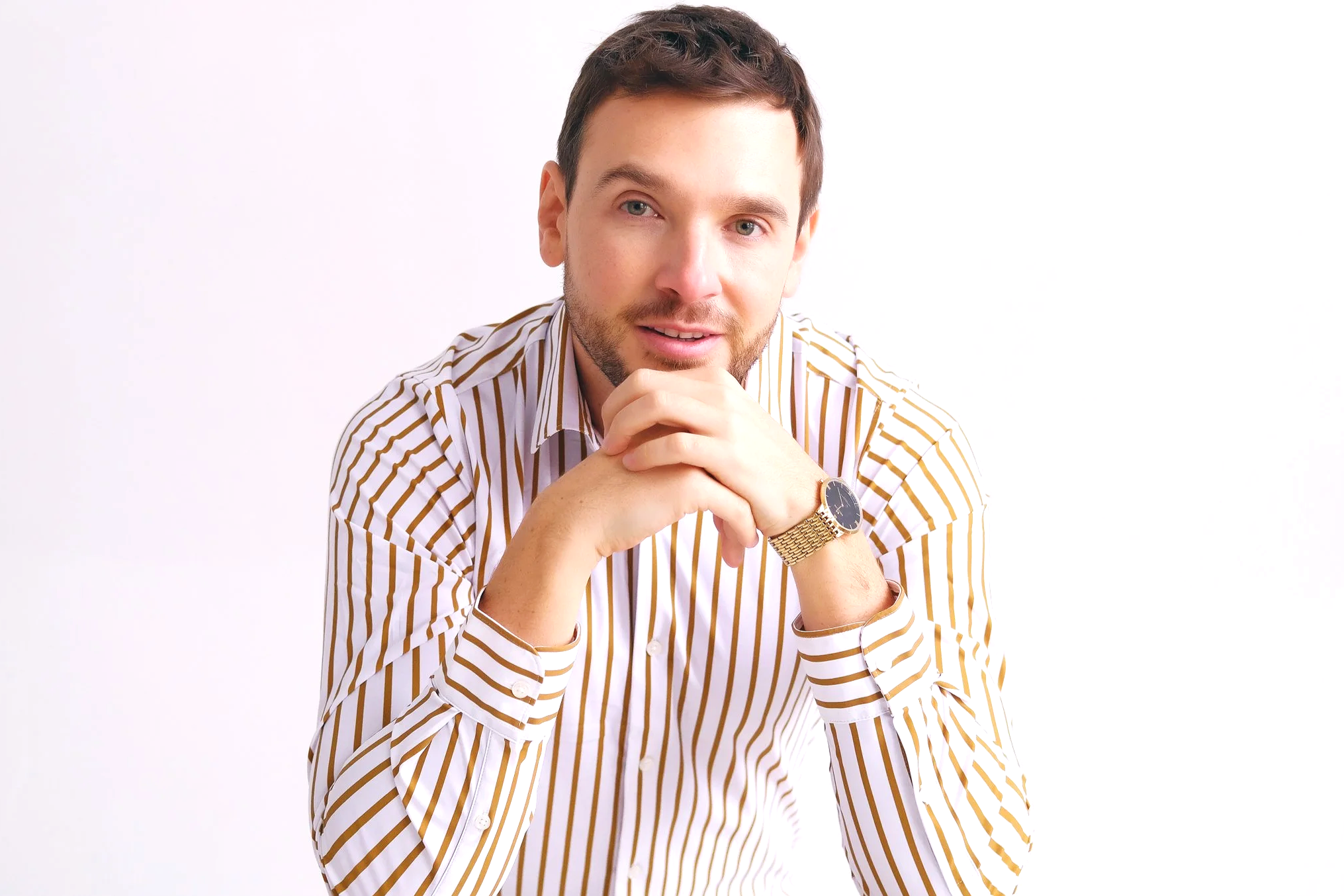 A man with short brown hair, wearing a striped shirt and a gold watch, resting his chin on his clasped hands, looking at the camera, against a plain white background.