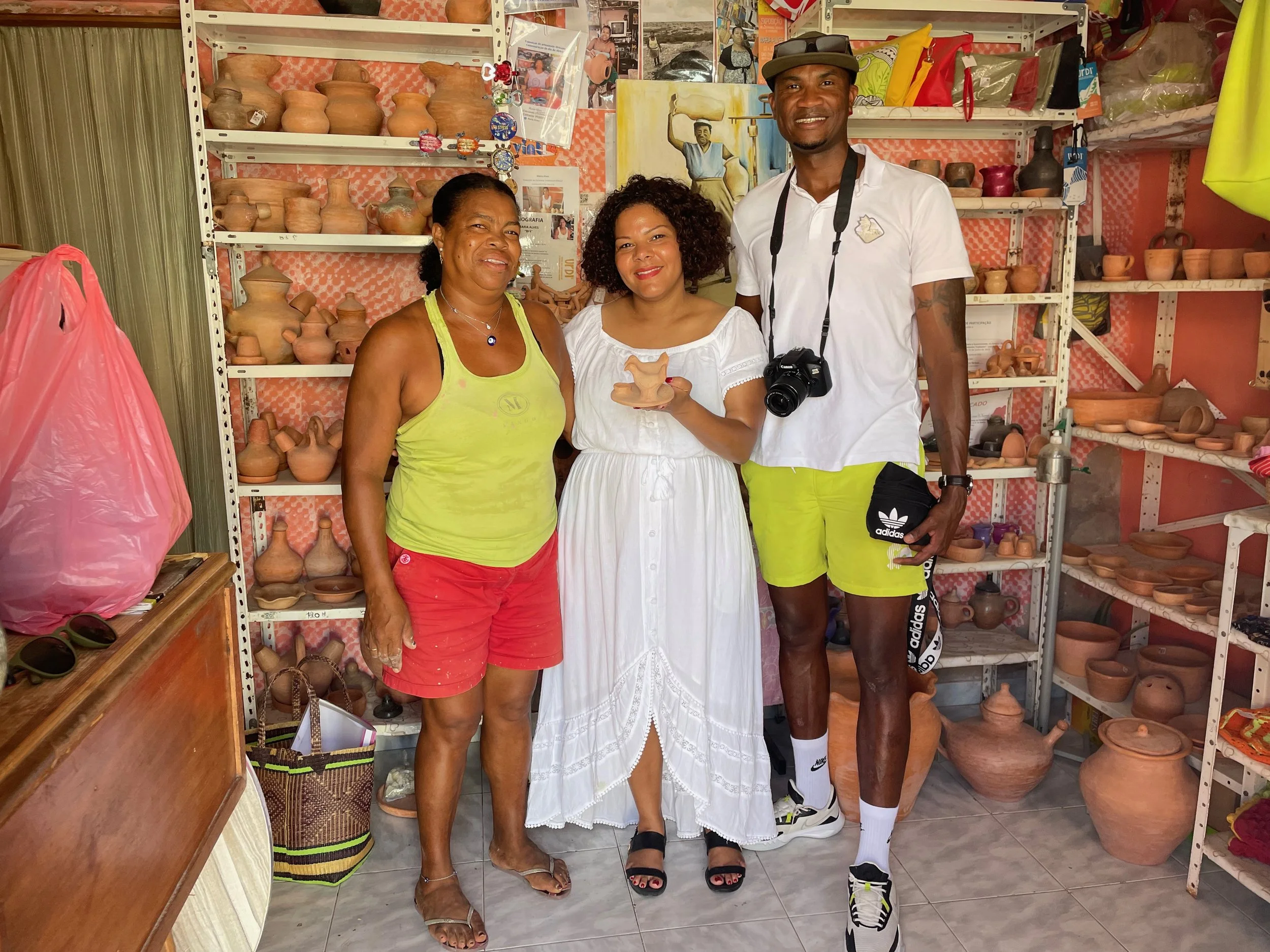 Three people standing in front of shelves filled with pottery in a shop or studio. The woman on the left is wearing a yellow tank top and red shorts, the woman in the middle is wearing a white dress and holding a small pottery piece, and the man on the right is wearing a white shirt, yellow shorts, and has a camera around his neck.