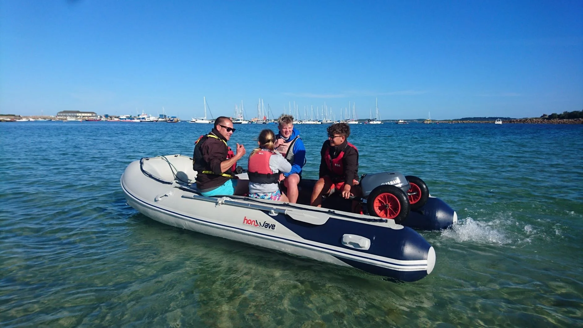 Four people on an inflatable boat with a motor, wearing life jackets, on a clear blue body of water with sailboats in the background.