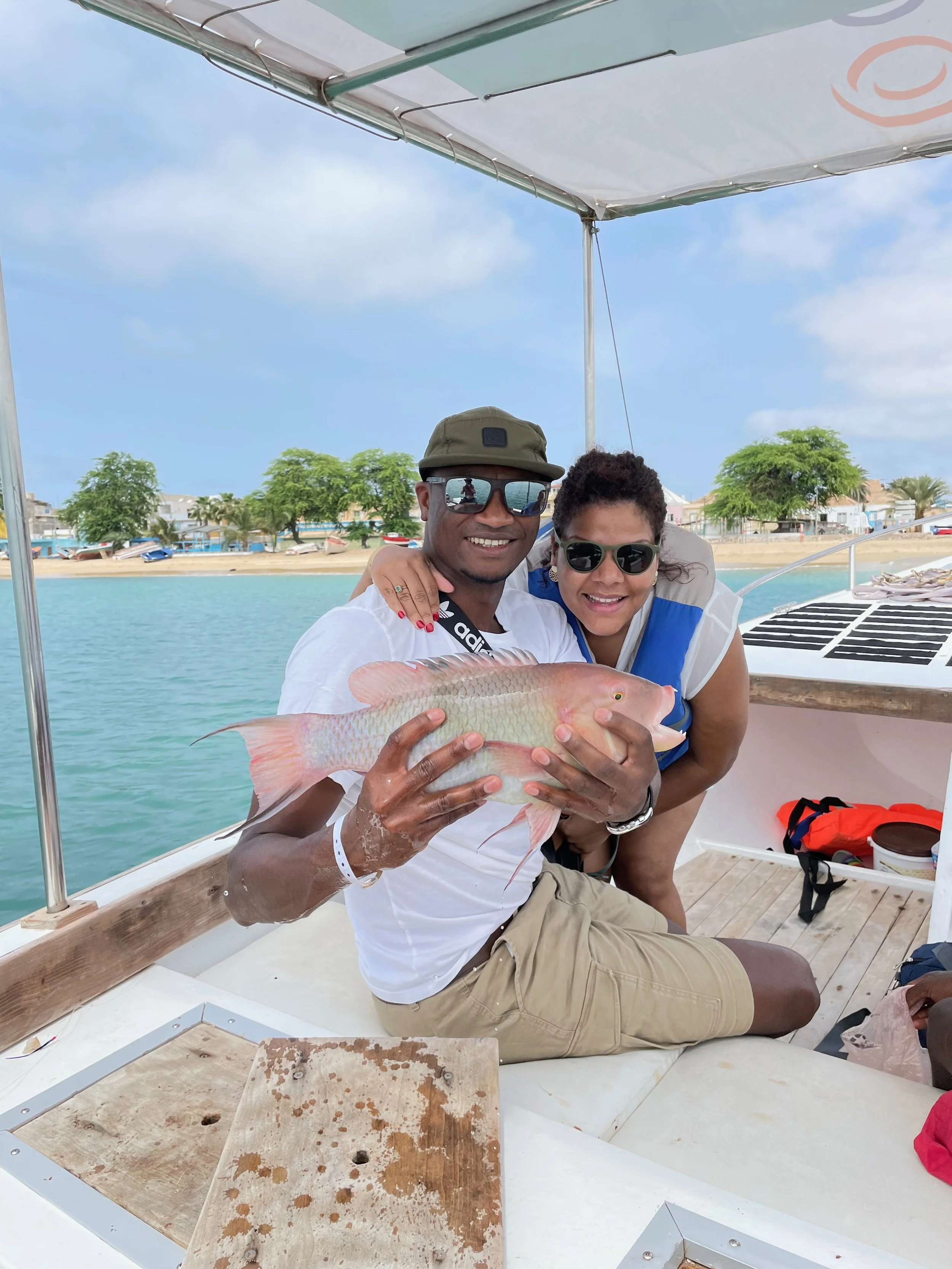 A man and woman on a boat holding a large fish, with a shoreline with trees and houses in the background, under a partly cloudy sky.