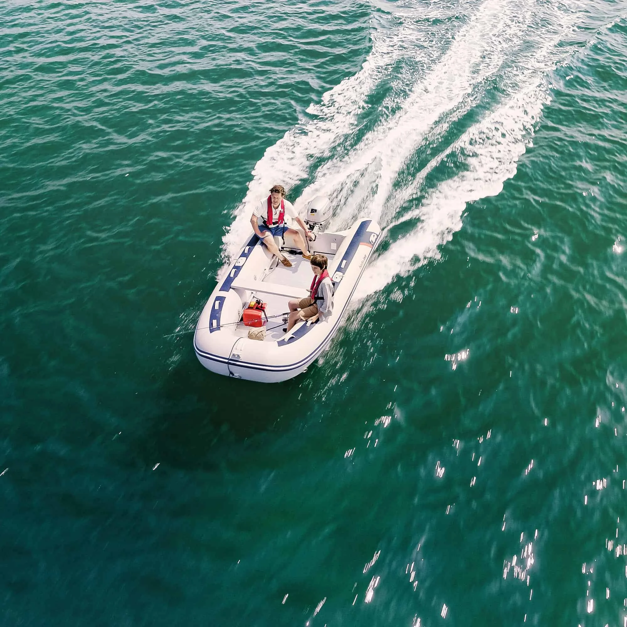 Two young men in casual clothing and life jackets riding a small inflatable boat with an outboard motor on the water.