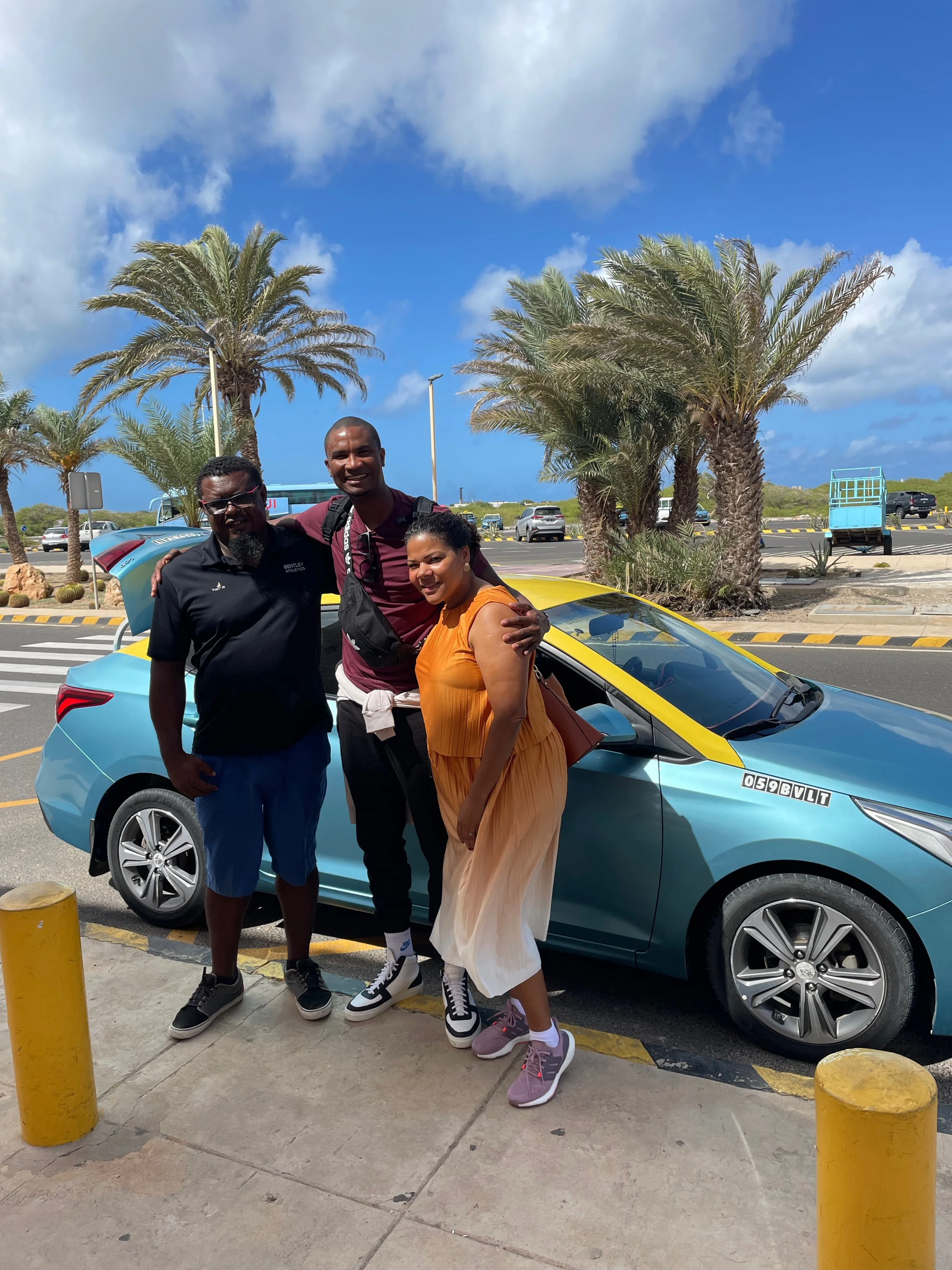 Three people smiling and standing together outdoors near a blue electric car with yellow accents, with palm trees and a parking lot in the background.