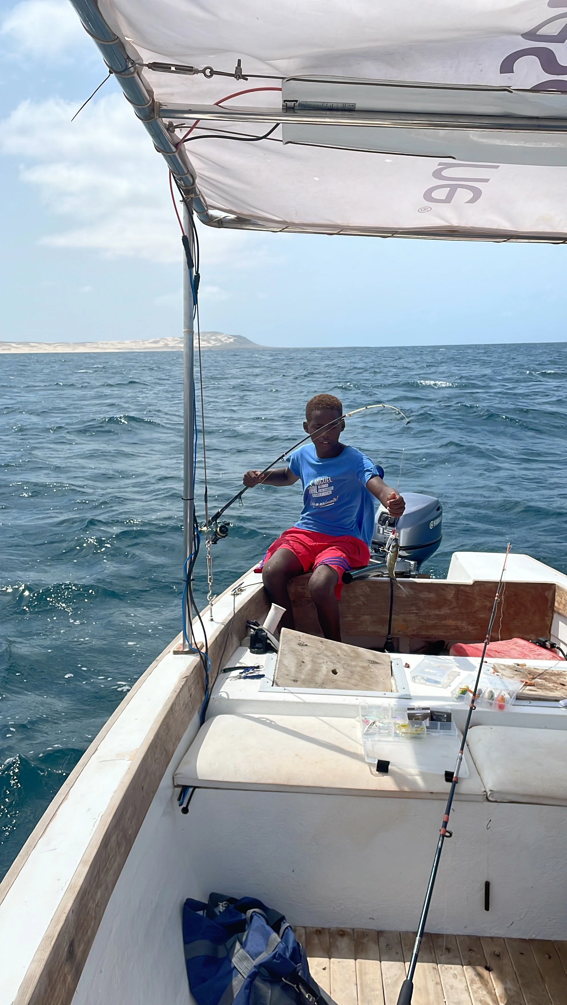 A boy sitting on a boat holding a fishing rod with a fish on the line, in the ocean with distant land and clouds in the background.