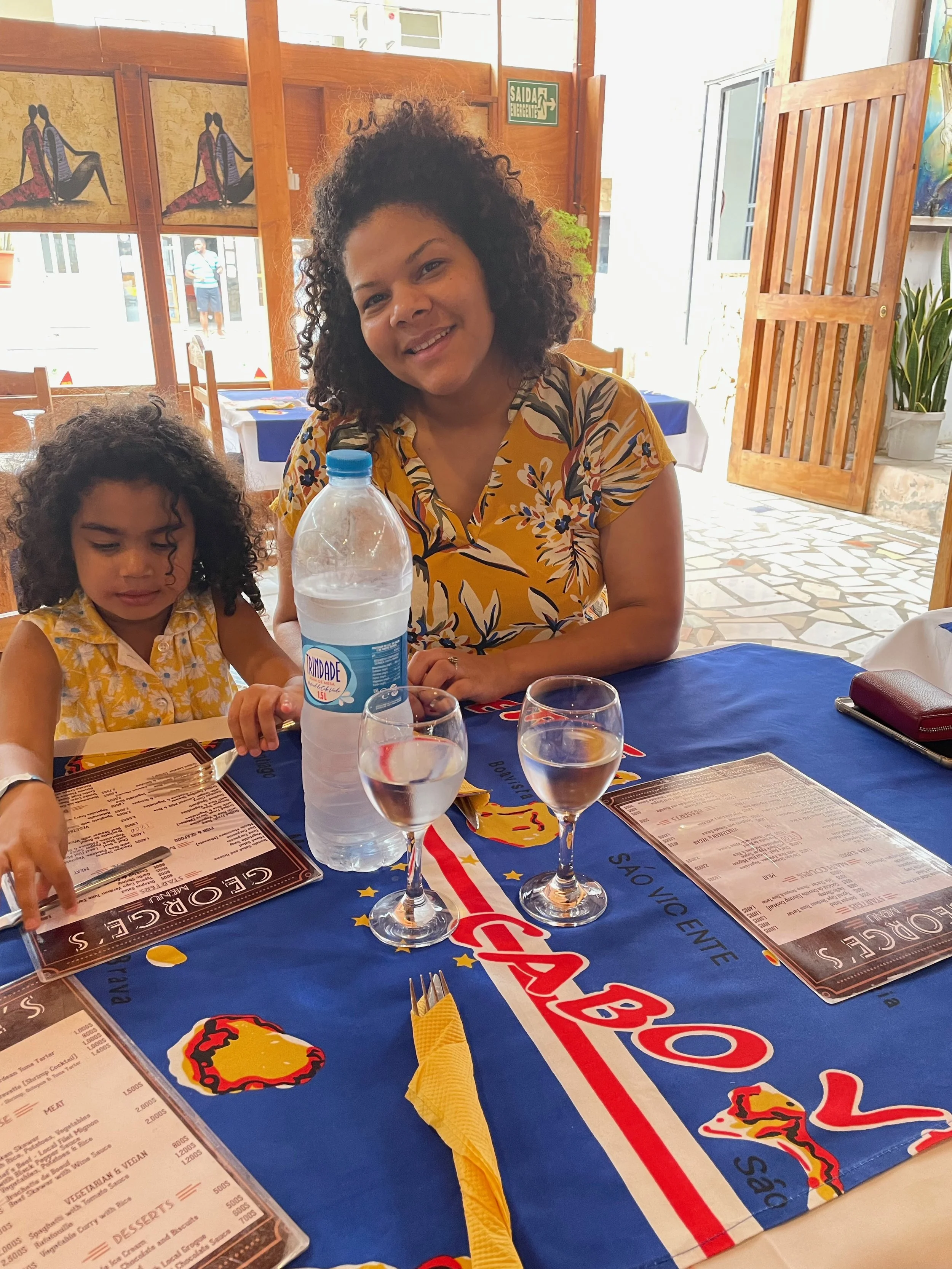 A woman and a young girl sitting at a restaurant table with a blue tablecloth that has the logo and text of a pizzeria. The woman is smiling and wearing a yellow floral dress, while the girl is looking down and wearing a yellow patterned dress. There are two glasses of water, a bottle of water, and menus on the table.
