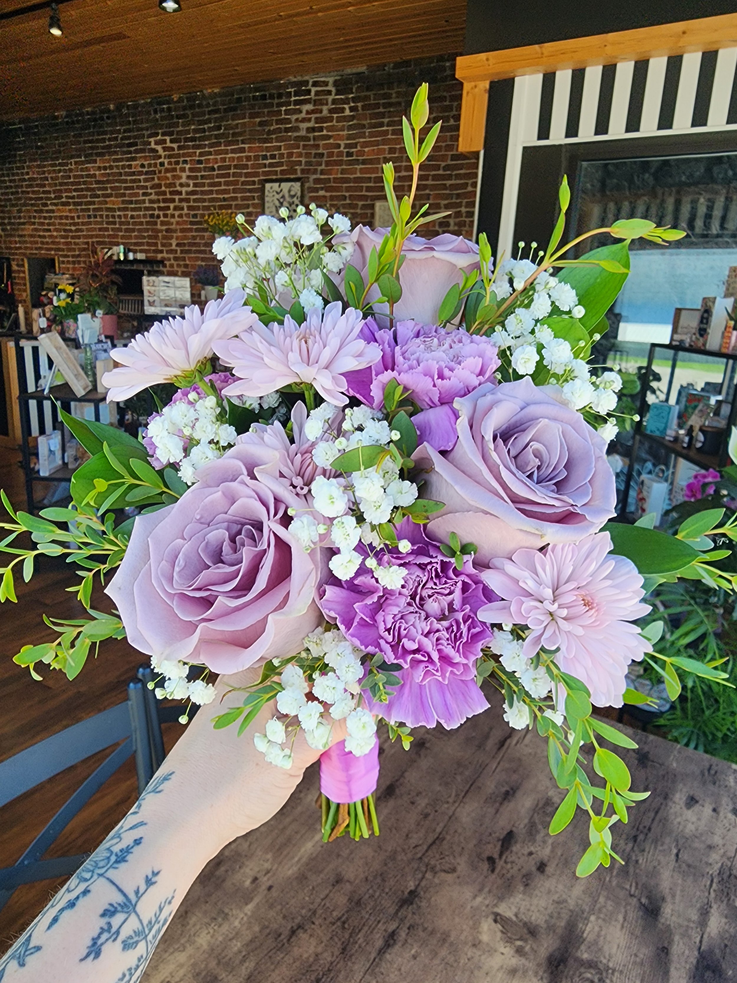A person holding a bouquet of purple and white flowers inside a room with a brick wall and wooden ceiling.