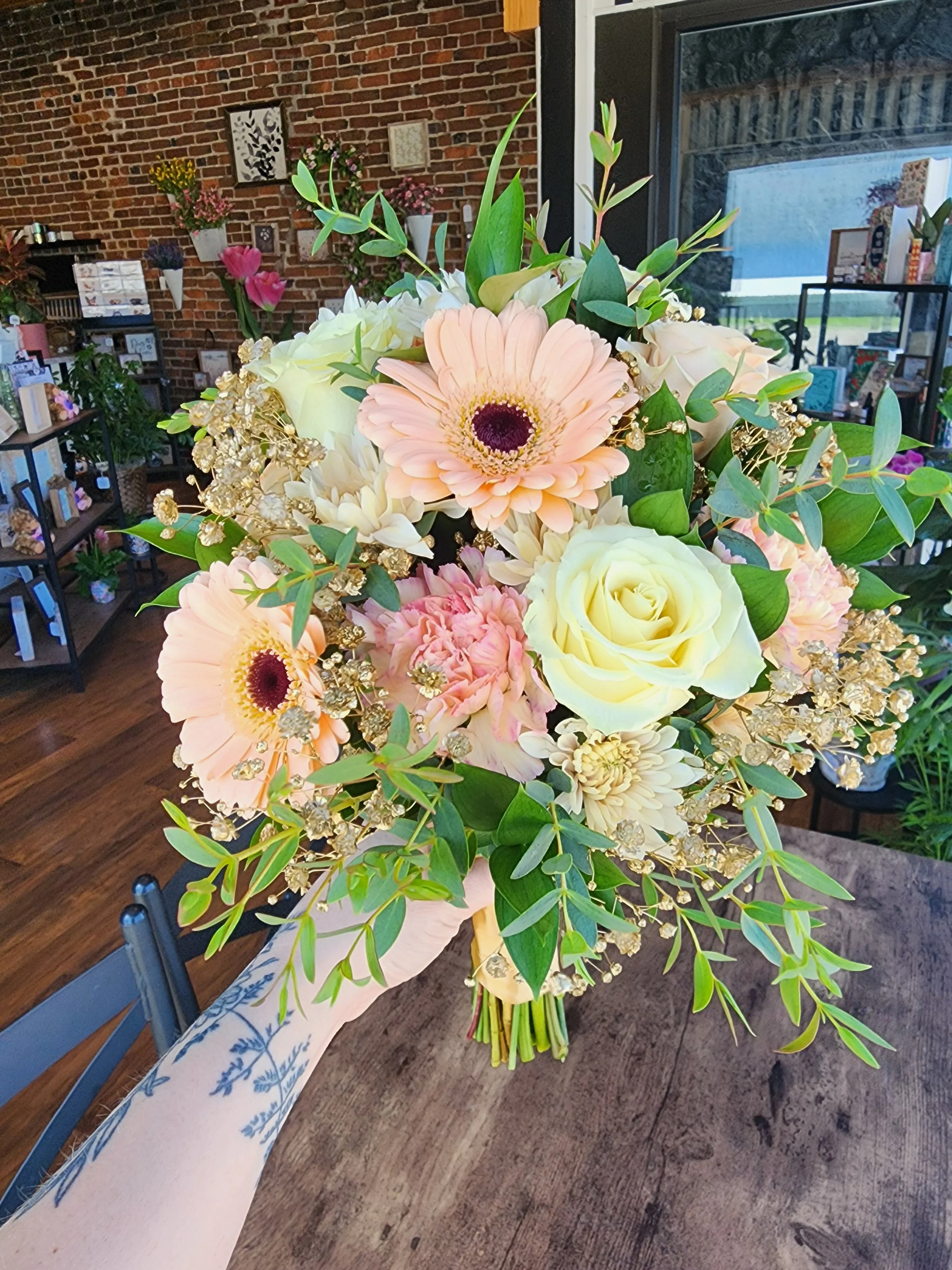 A large bouquet of white, pink, and yellow flowers with green leaves on a table in a room with brick wall, shelves with flowers, and a window.