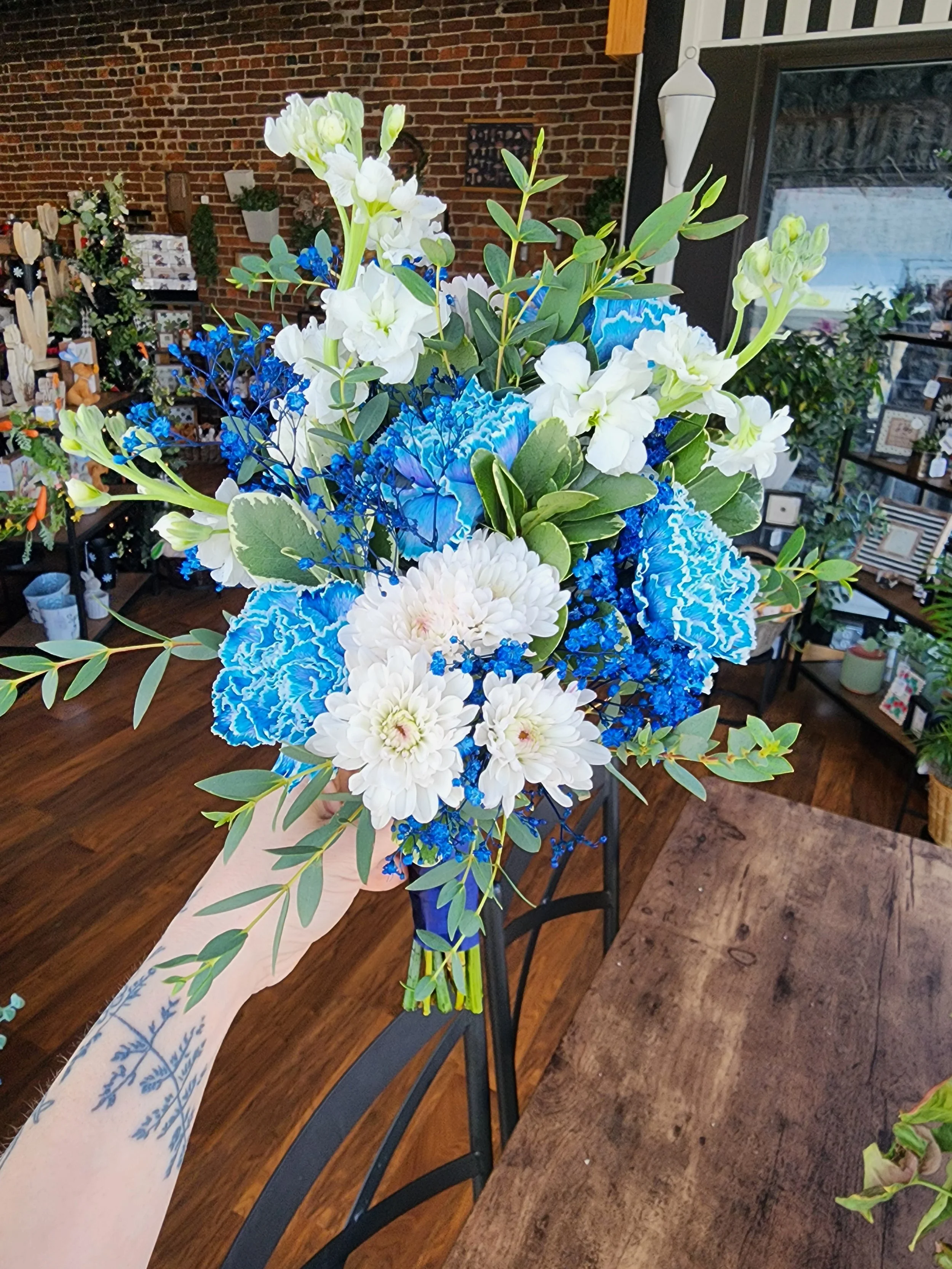 A vibrant flower arrangement with white delphiniums, white chrysanthemums, and blue carnations, accented by green foliage, held in hand, against a background of a retail or gift shop with brick wall and shelves of decorative items.