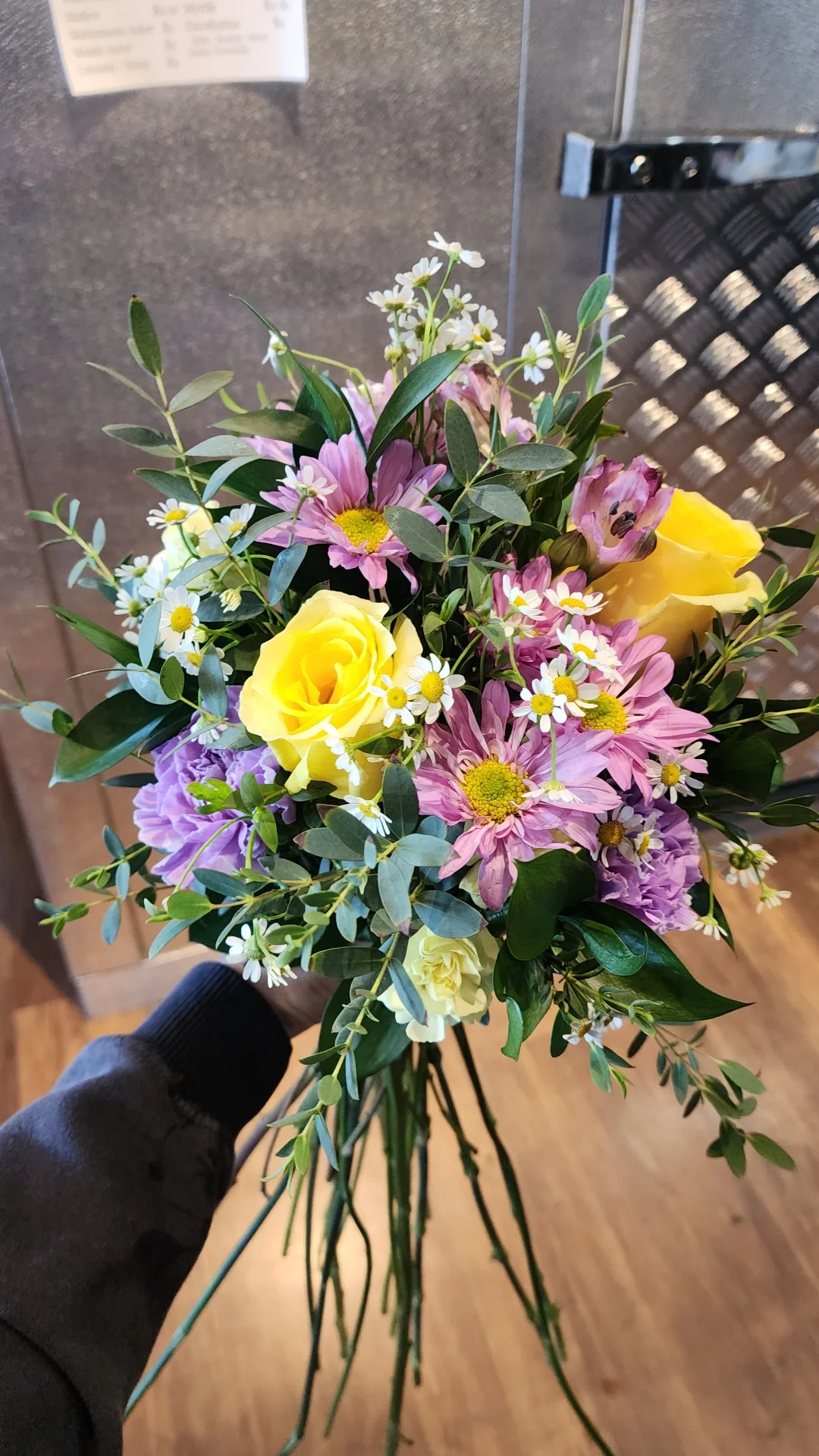 A hand holding a bouquet of mixed flowers, including yellow roses, pink daisies, white daisies, and greenery, with a metallic background.
