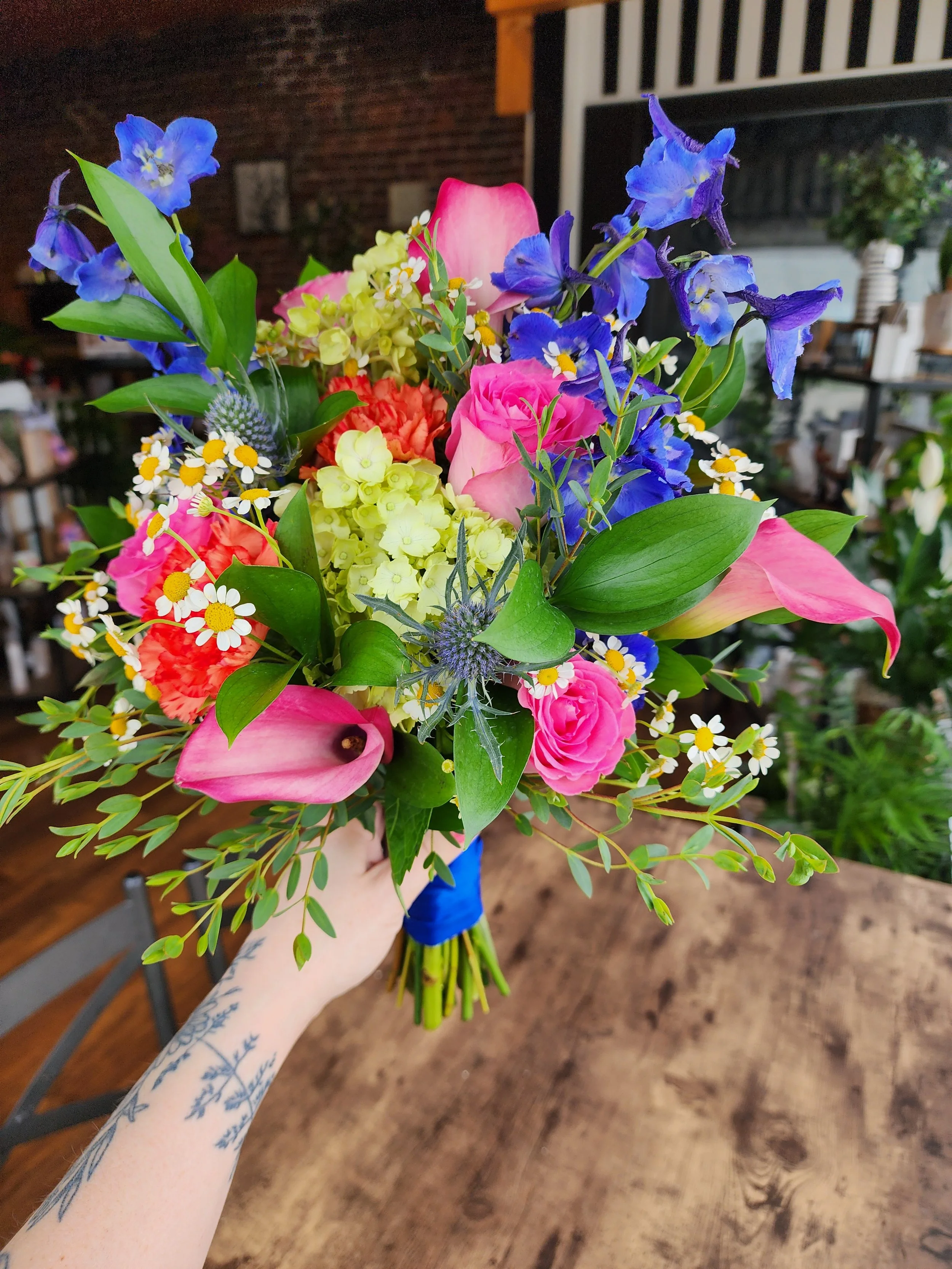 Close-up of a colorful bouquet of flowers held by a person's hand with a tattoo on their forearm, in a room with wooden flooring and furniture in the background.