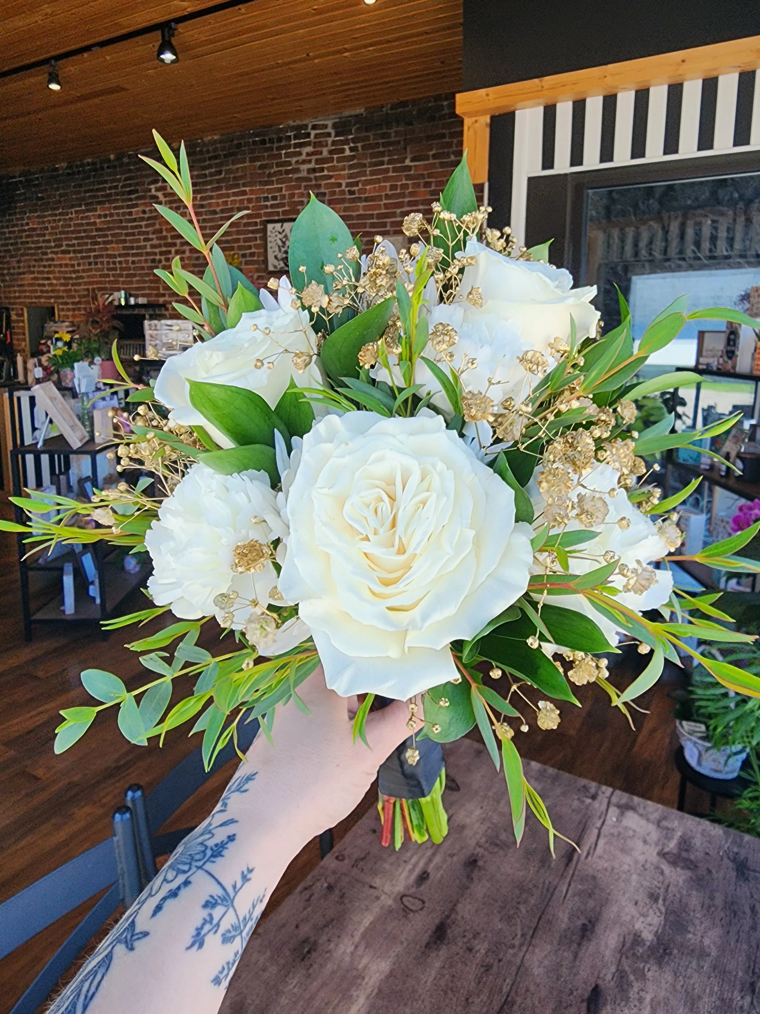 Hand holding a bouquet of white roses, greenery, and gold accents in a room with brick wall, wooden ceiling, and television.