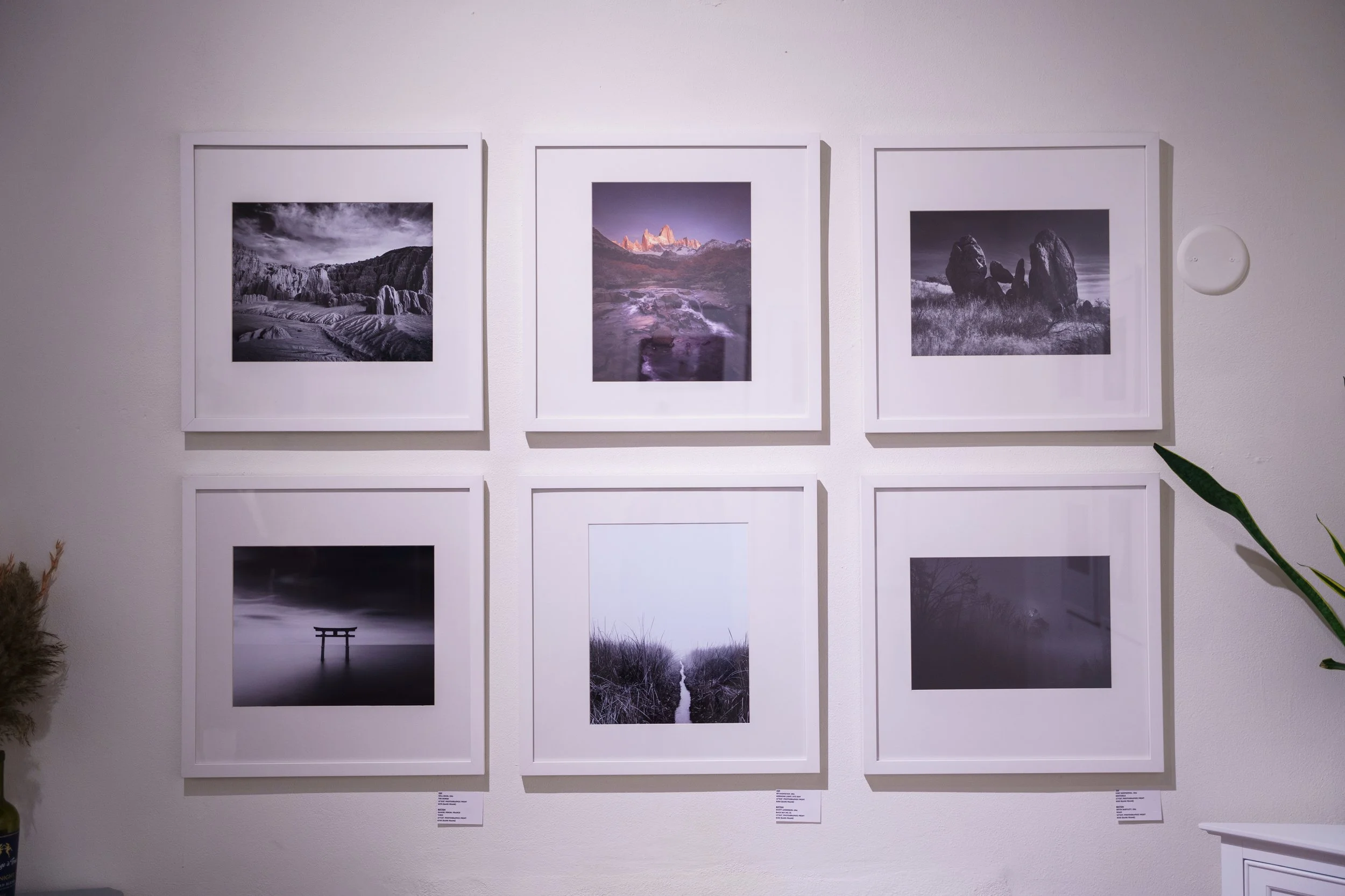 Gallery wall displaying six framed black-and-white landscape photographs, including mountains, water, rocks, and natural scenery, arranged in two rows on a white wall.