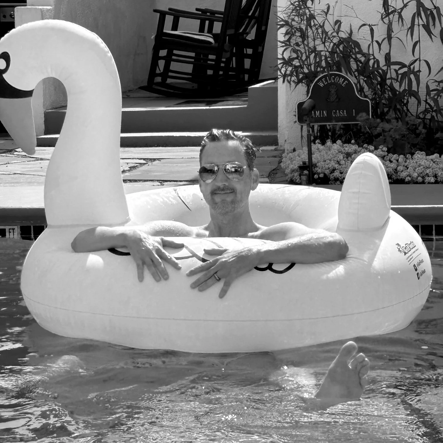 A man relaxing in an inflatable swan-shaped pool float, wearing sunglasses, with a rocking chair and house sign in the background.