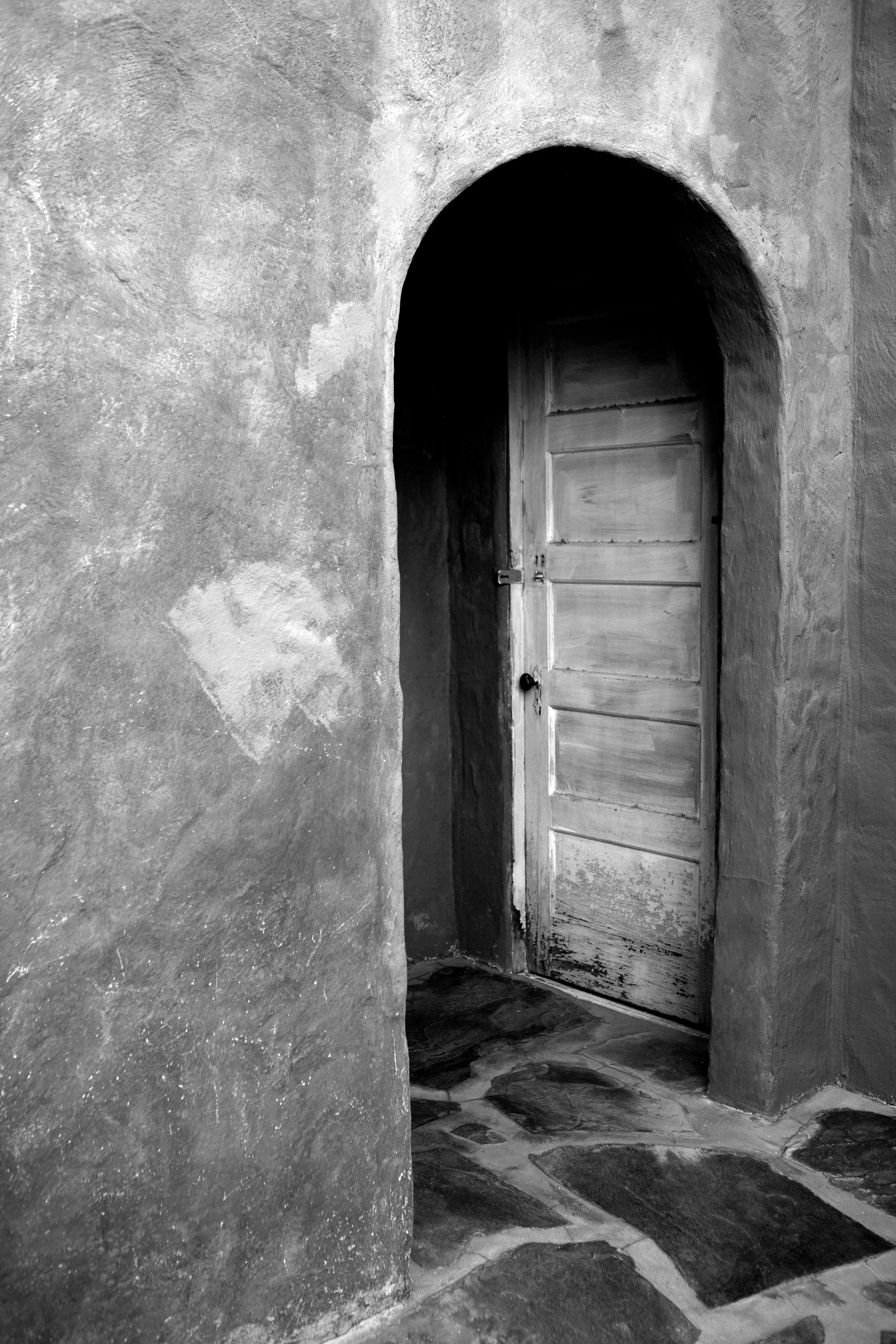 Black and white photo of an old wooden door in an arched passageway with textured stone walls and a patterned stone floor.