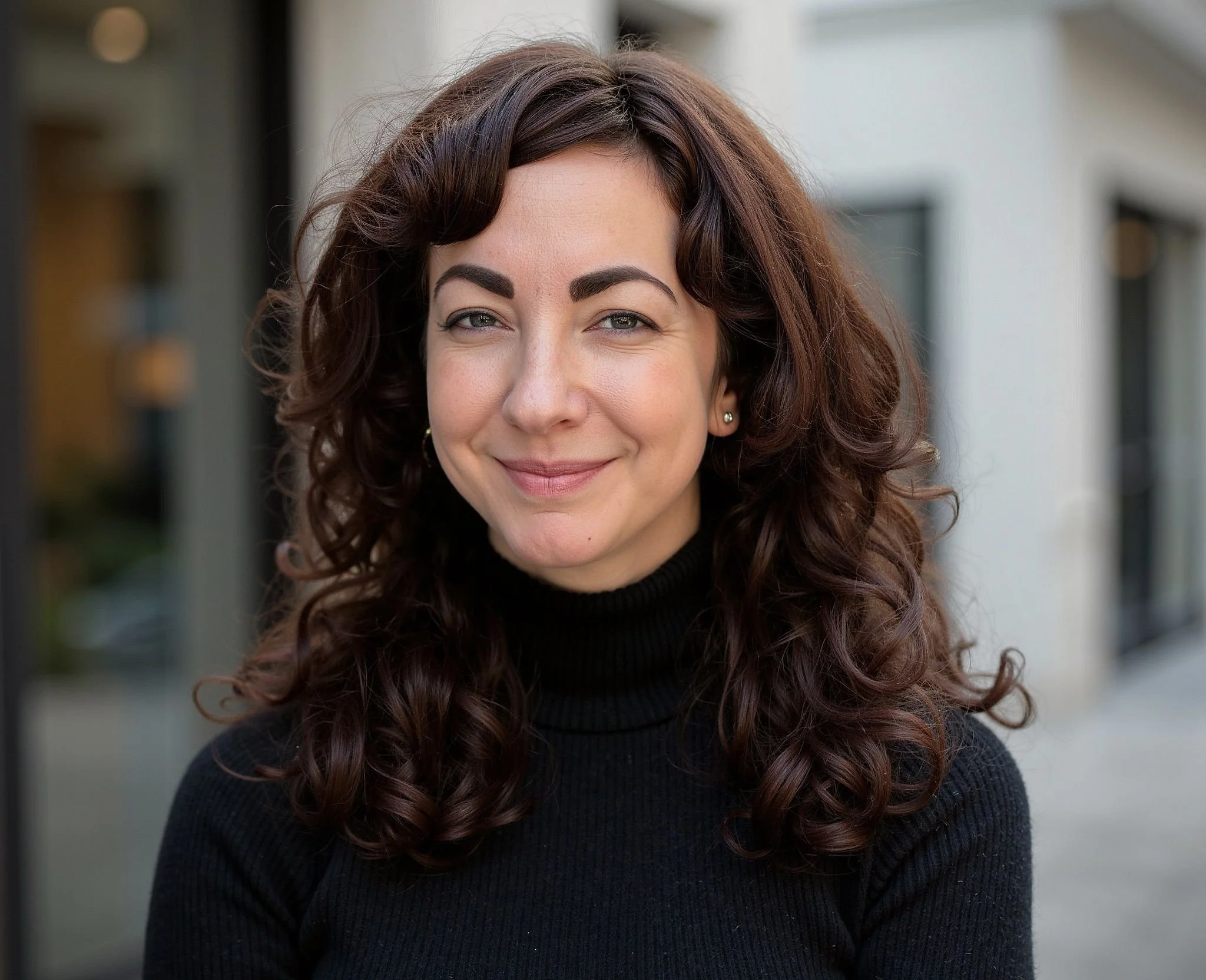 A woman with dark brown, curly hair smiling outdoors, wearing a black turtleneck sweater.